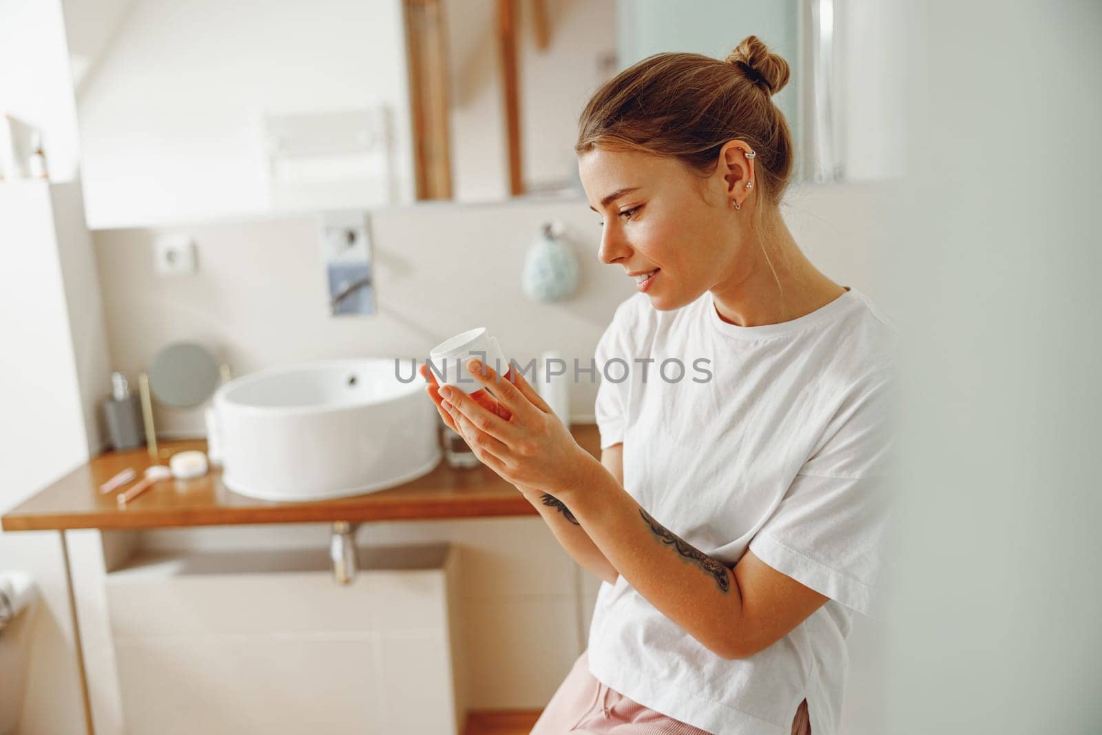 Young woman looking on jar with skincare cream sitting in bathroom. Home beauty routine by Yaroslav_astakhov