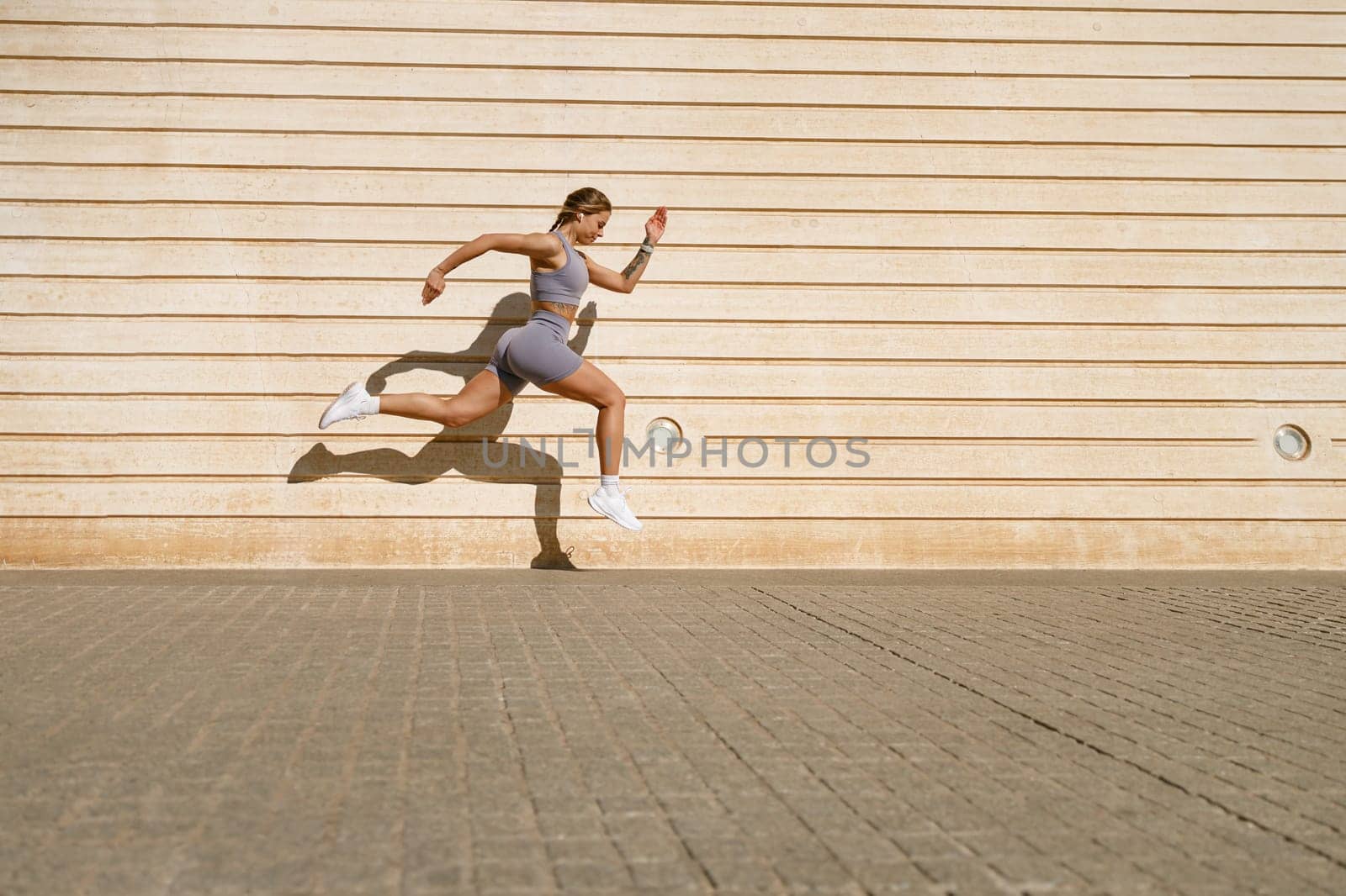 Female athlete in sportswear leaping in air with wall backdrop outdoors. Healthy lifestyle concept by Yaroslav_astakhov