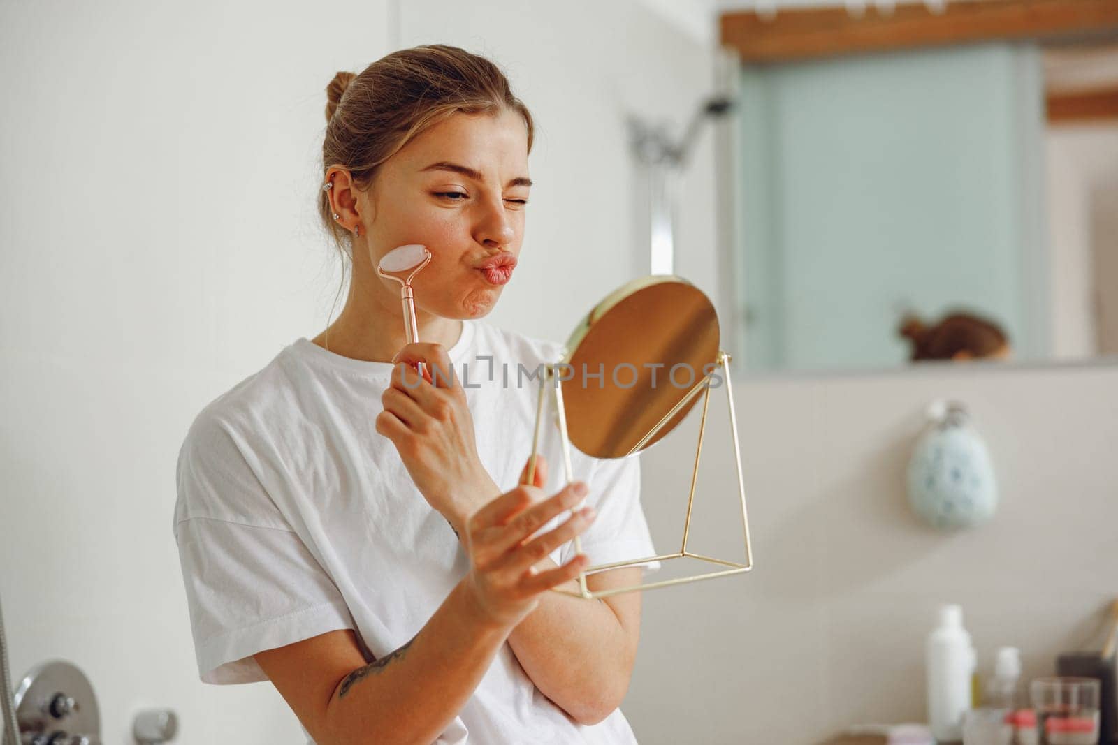 Smiling woman massaging her face with jade roller massager and looking at round cosmetic mirror