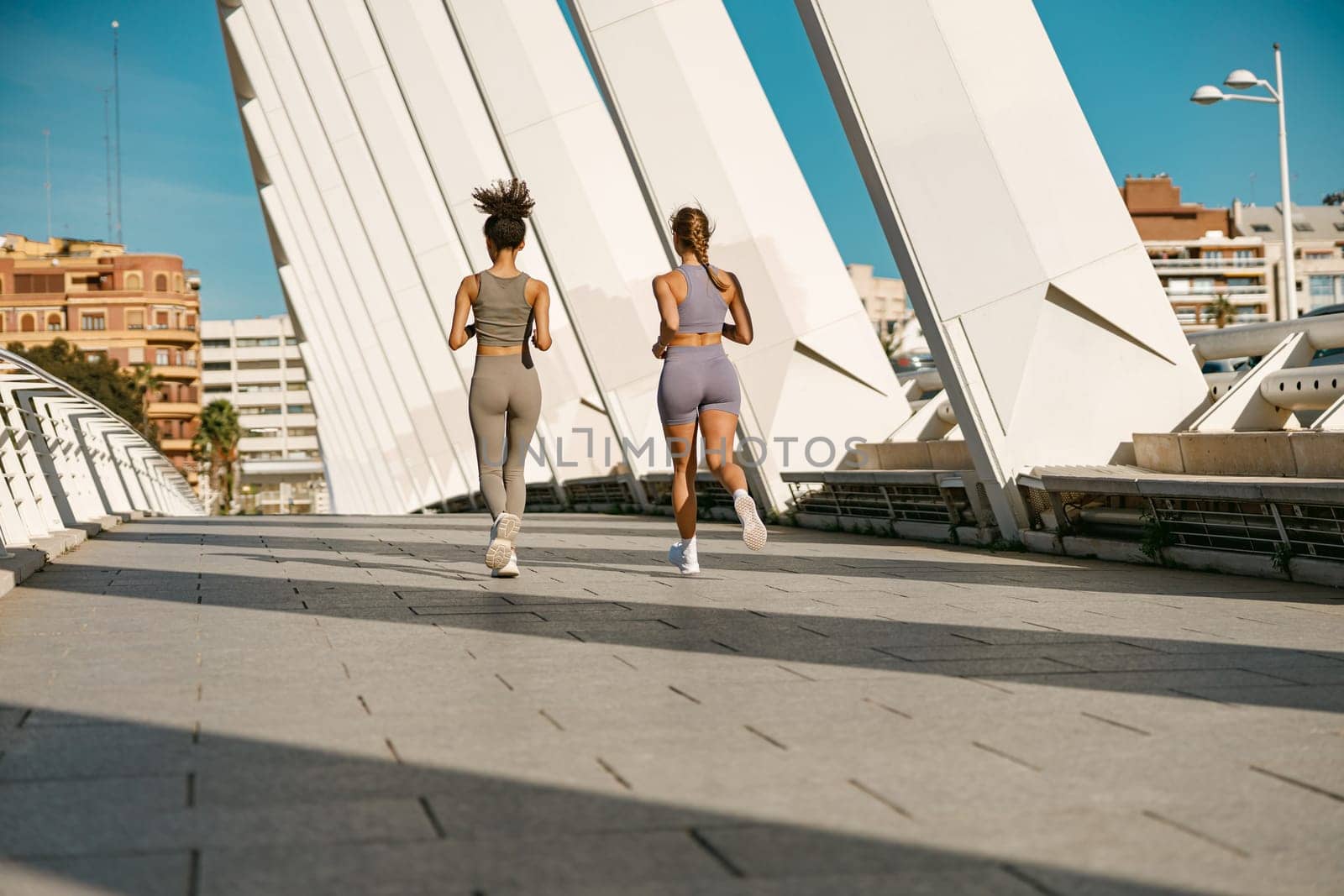 Back view of two women athlete running side by side along an outdoor track on buildings background by Yaroslav_astakhov