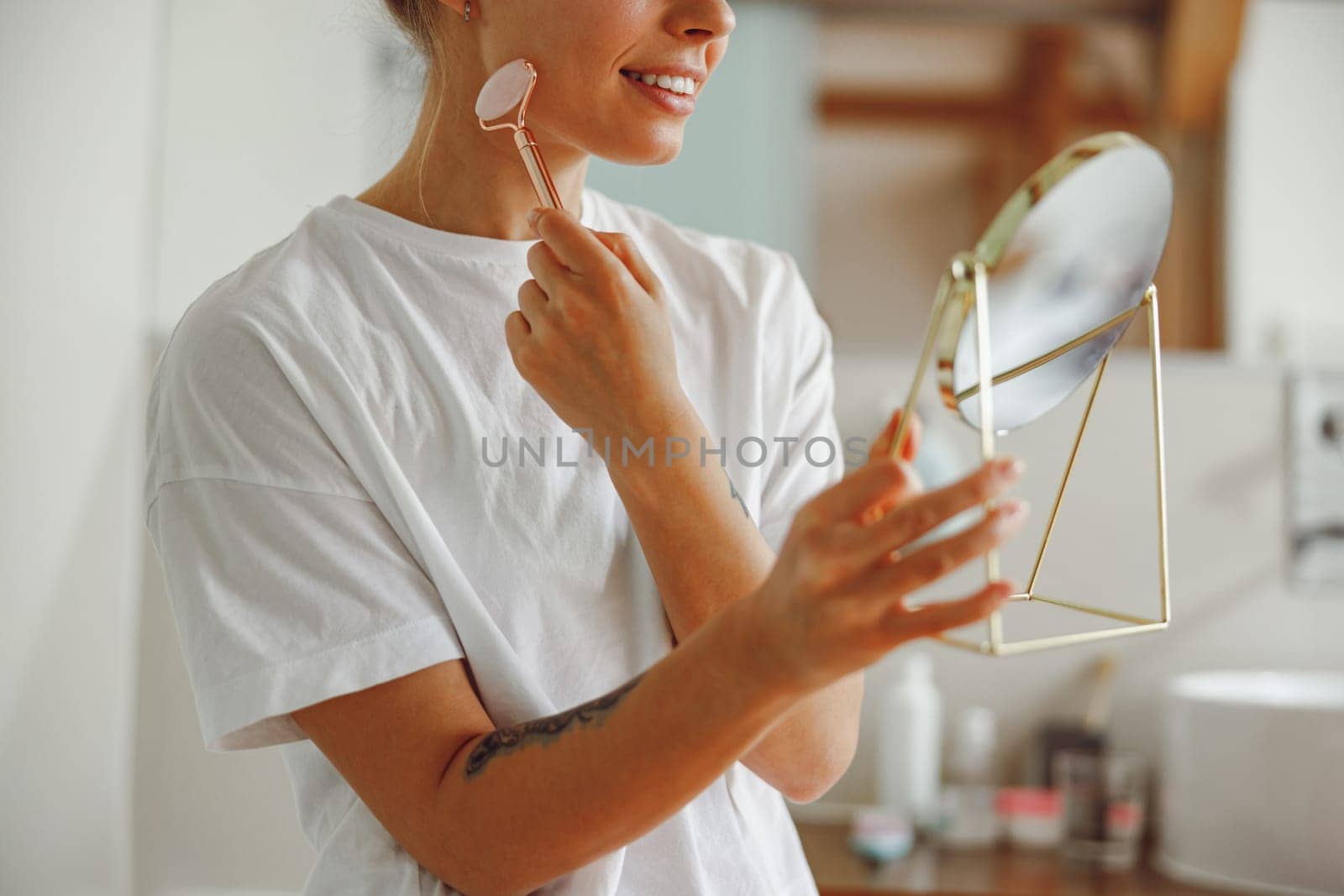 Close up of woman massaging her face with jade roller massager and looking at round cosmetic mirror by Yaroslav_astakhov