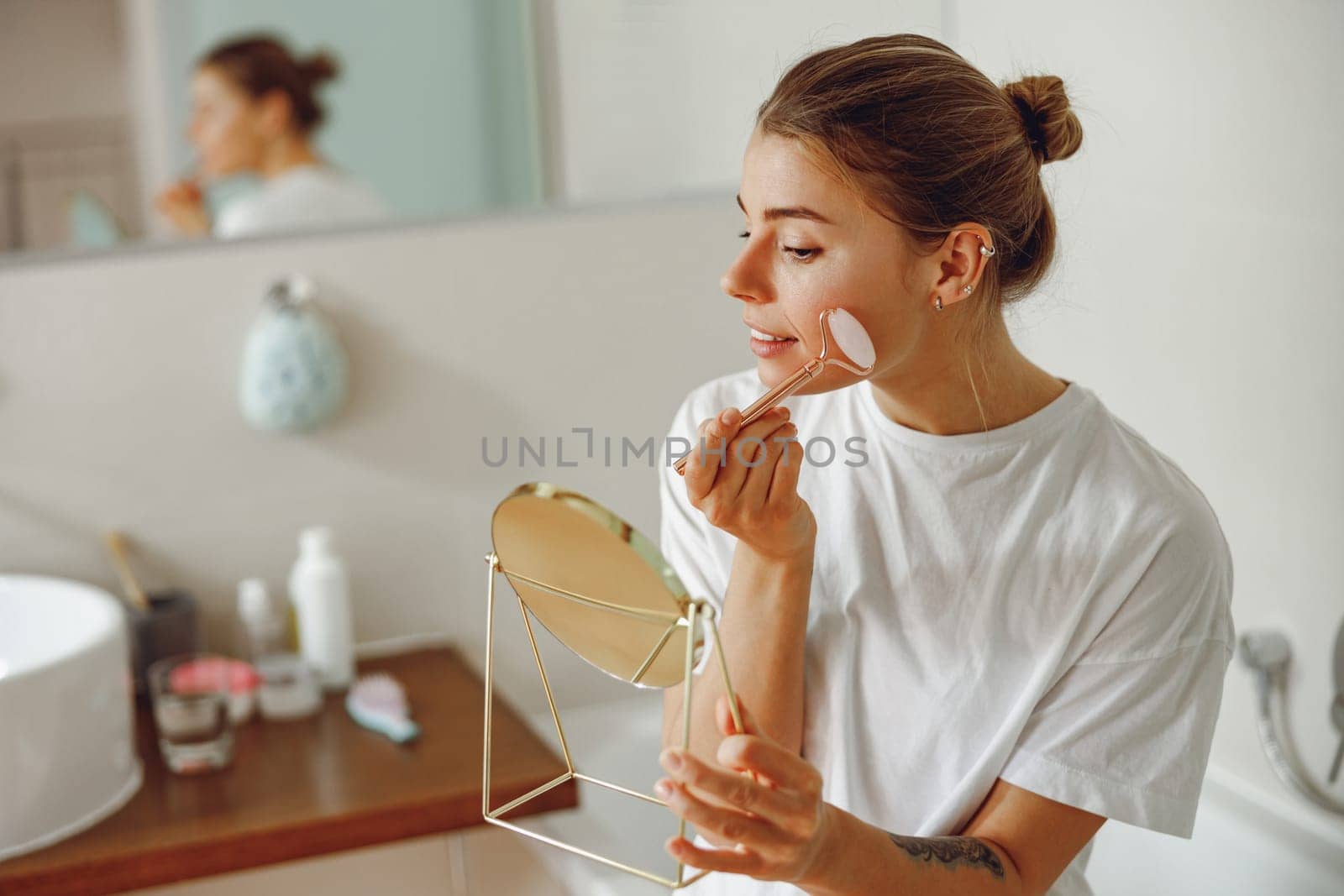 Young woman massaging face with jade roller and looking at round cosmetic mirror in bathroom by Yaroslav_astakhov
