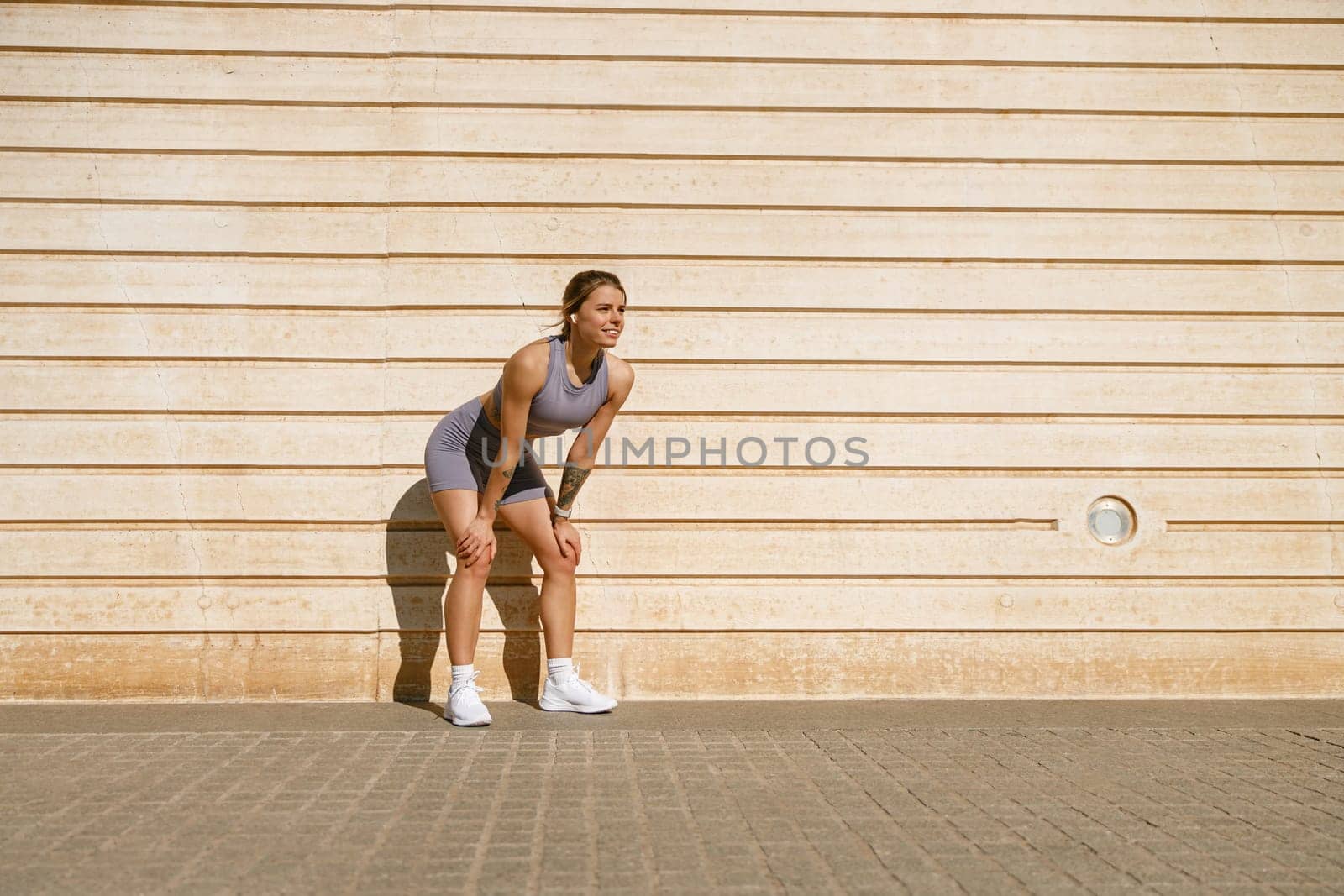 Smiling woman in sportswear have a rest after workout outside standing on wall background by Yaroslav_astakhov