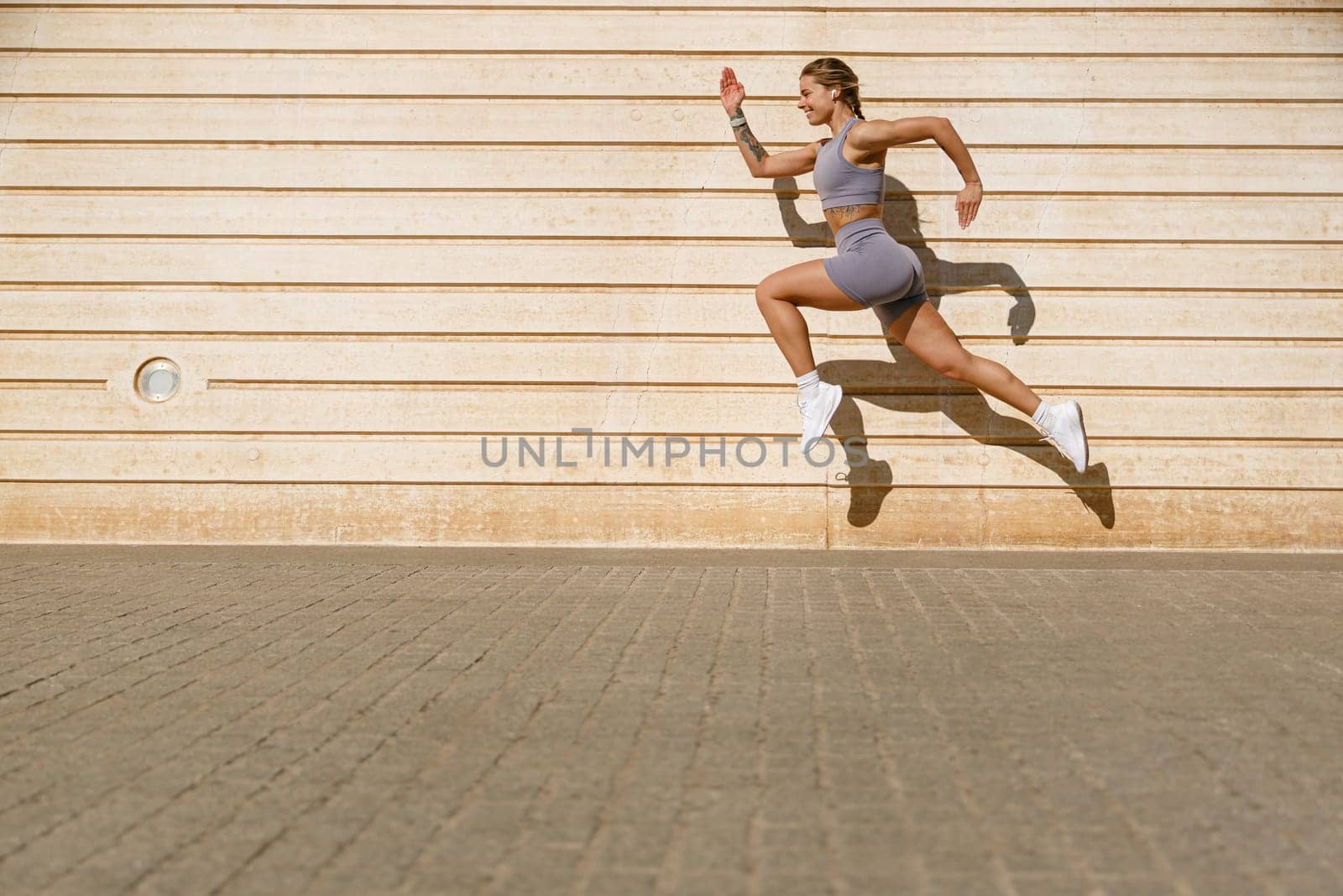 Female athlete in sportswear leaping in air with wall backdrop outdoors. Healthy lifestyle concept by Yaroslav_astakhov