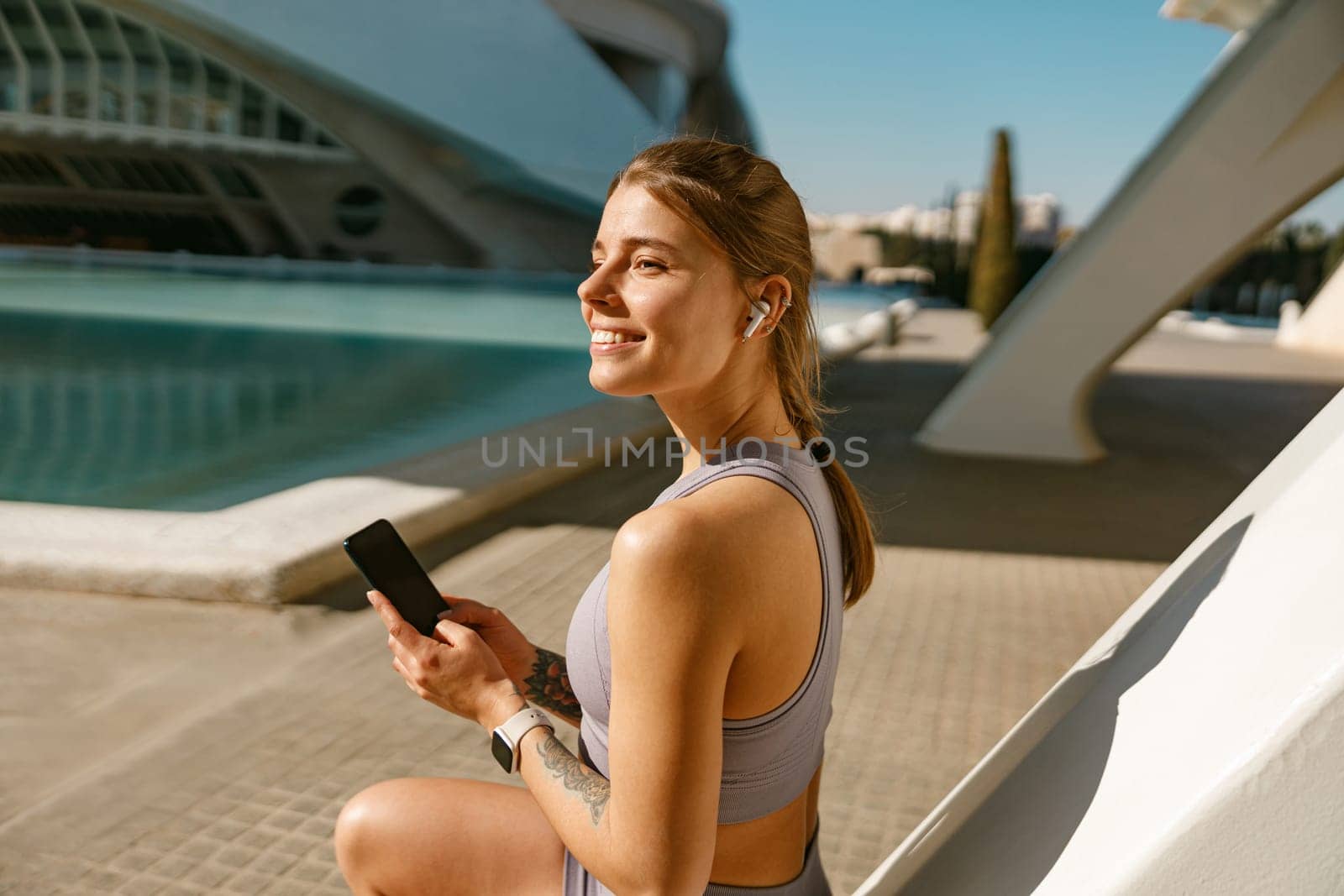 A young woman in sportswear is resting after a workout outside while standing with smartphone by Yaroslav_astakhov