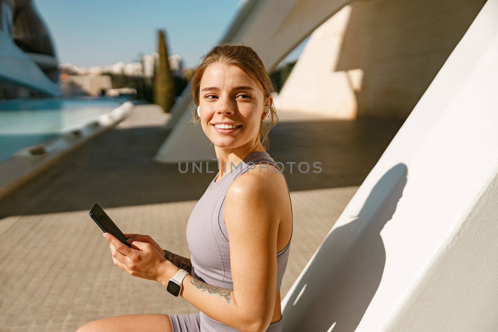 A young woman in sportswear is resting after a workout outside while standing with smartphone