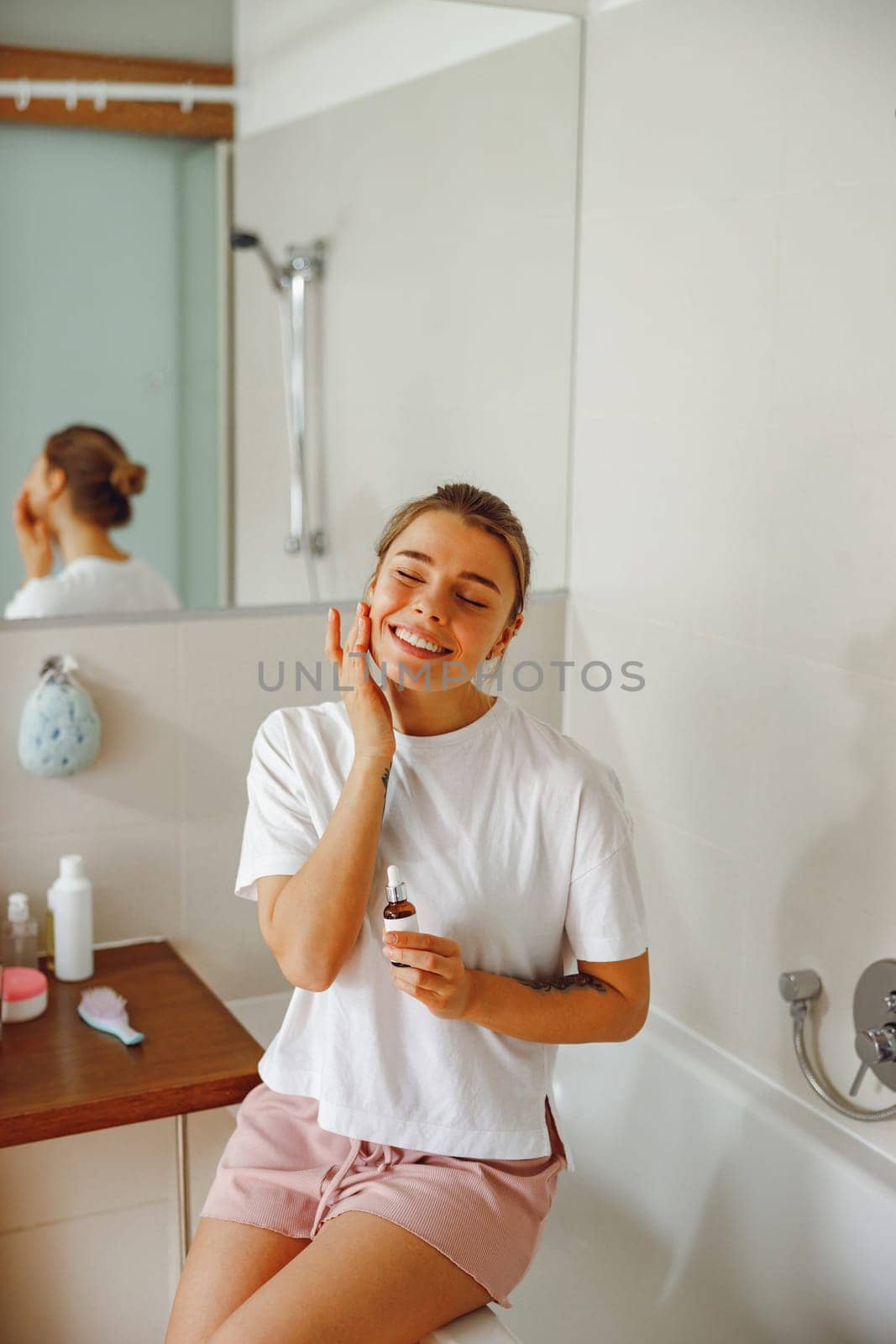 Smiling female applying serum on face moisturizing and caring for skin standing in bathroom by Yaroslav_astakhov
