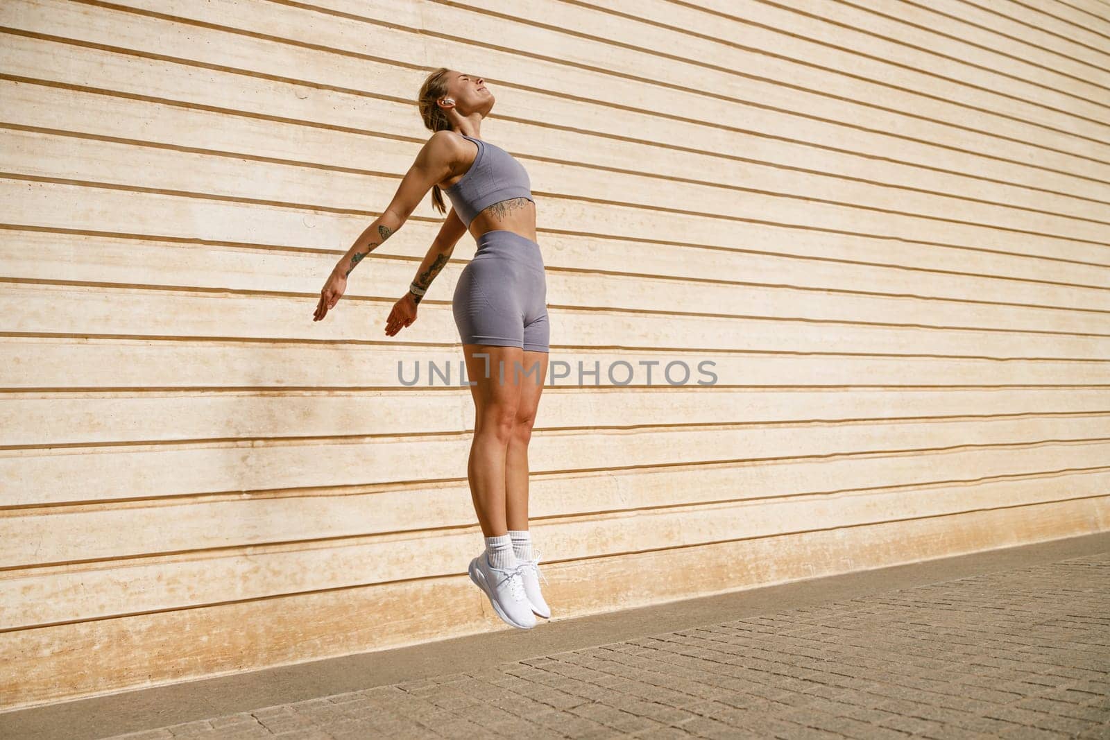 Female athlete in sportswear leaping in air with wall backdrop outdoors. Healthy lifestyle concept by Yaroslav_astakhov