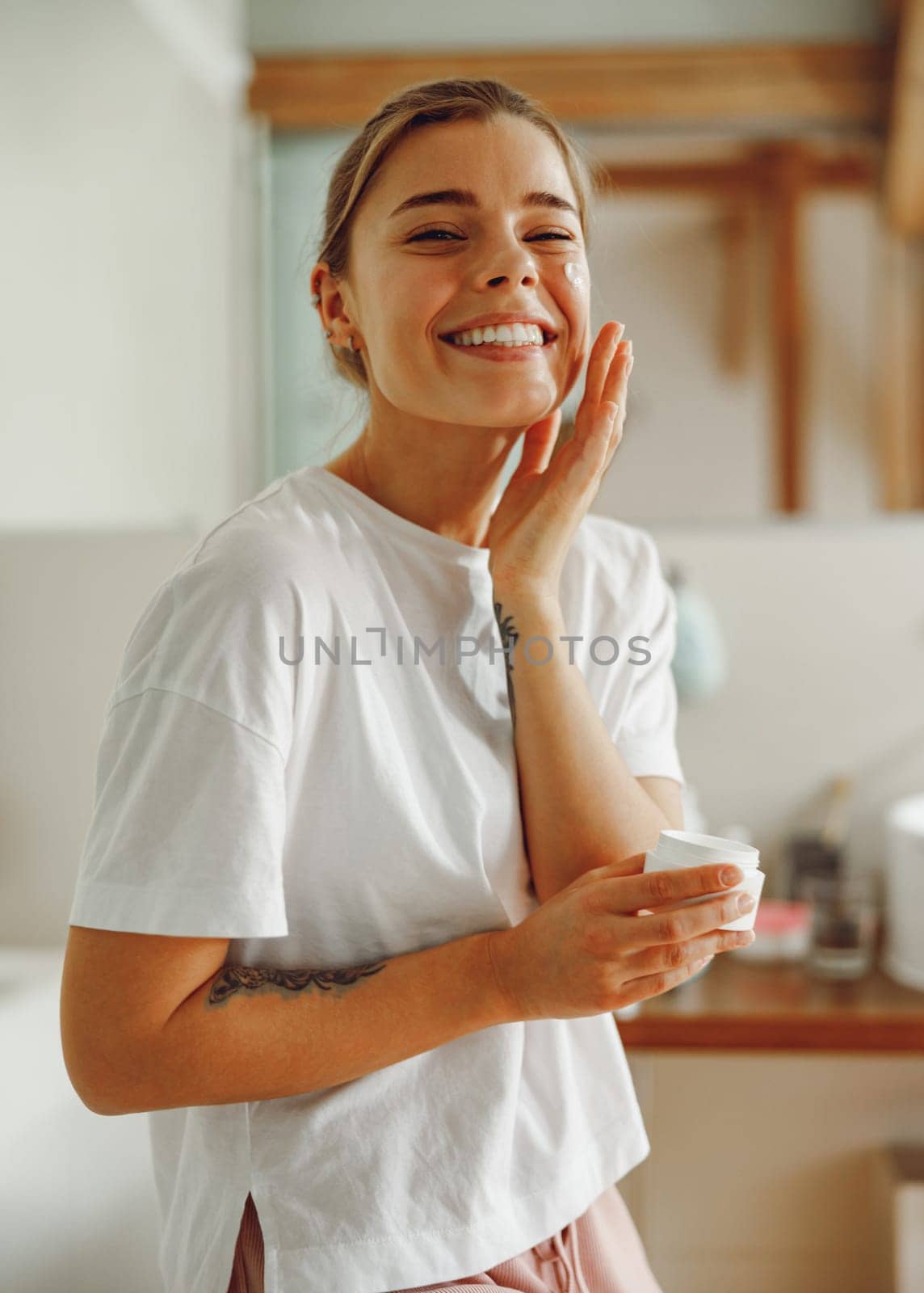 Happy woman applying hydrating moisturizer on her face sitting in bathroom. Home beauty routine