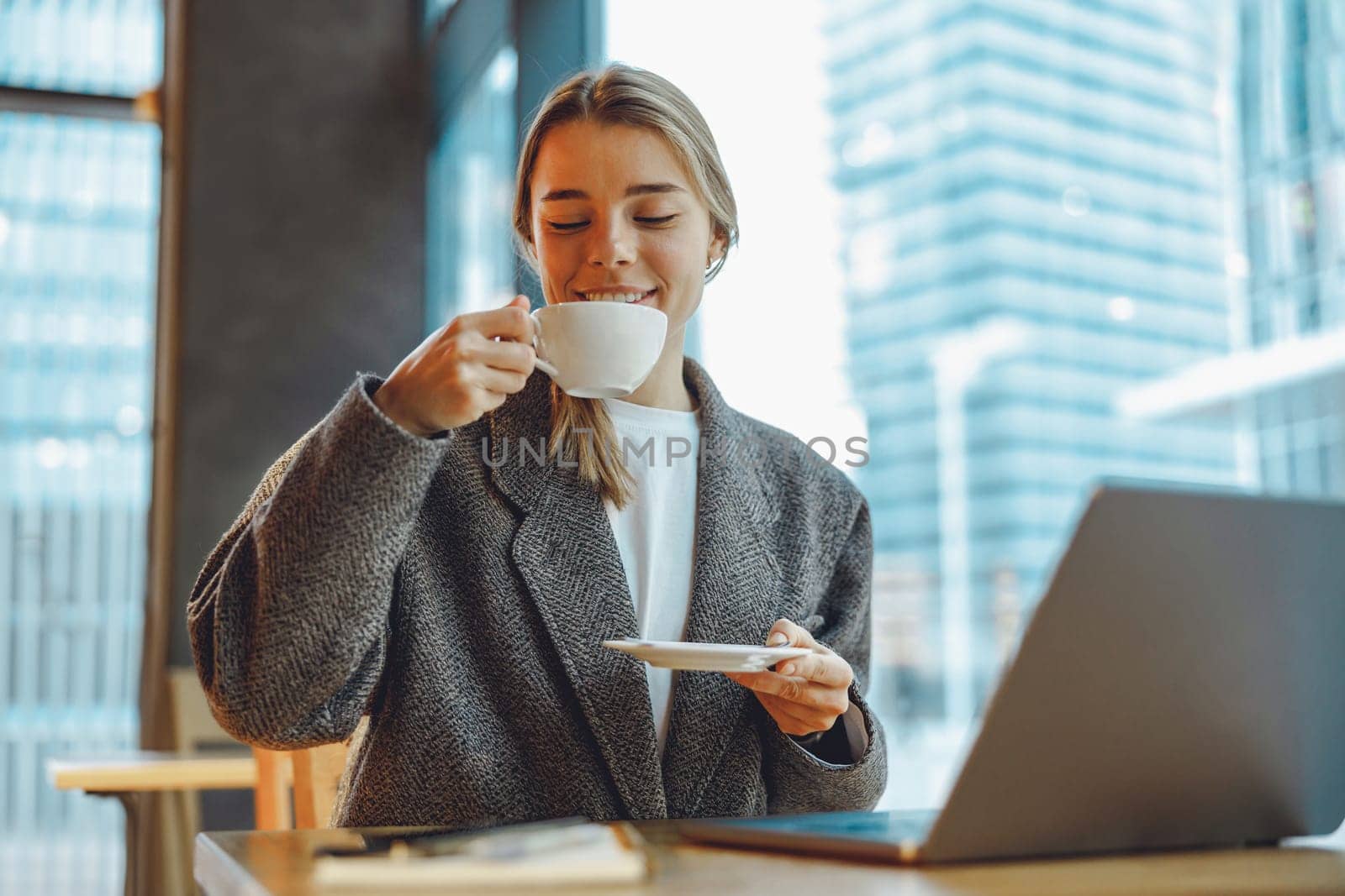 Smiling female entrepreneur is drinking coffee in coworking while working on laptop near window