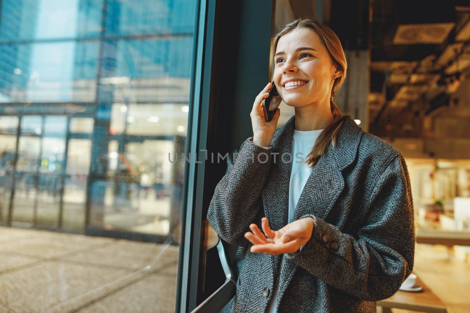 Stylish young female freelancer is talking phone with client while standing in coworking near window