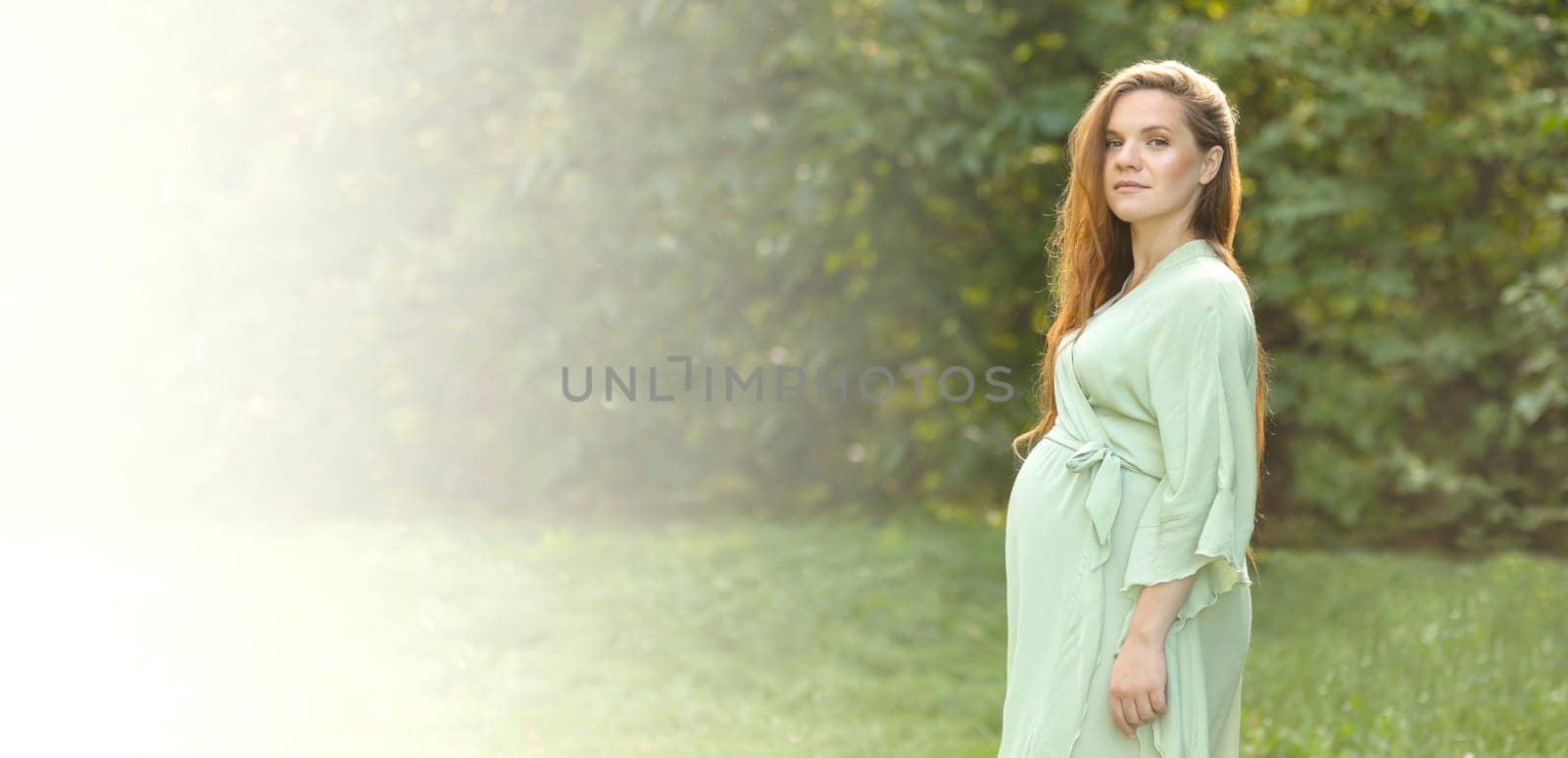 Banner white pretty pregnant woman stands, looks at camera. Green trees, park is on background. Long brown hair young healthy expectant wears dress. Baby shower, delivery,preparation concept.Copyspace
