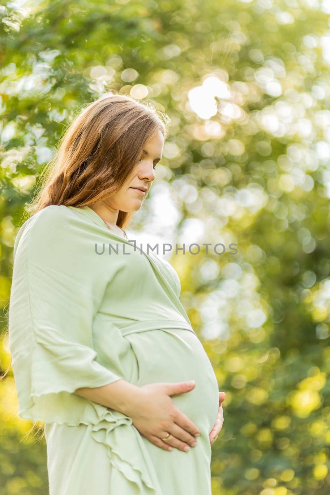 Young pregnant woman holds big belly, green trees,sky on background. Brown long haired female wear dress. Magic happy pregnancy. Child delivery preparations,emotional connection with baby. Vertical by netatsi