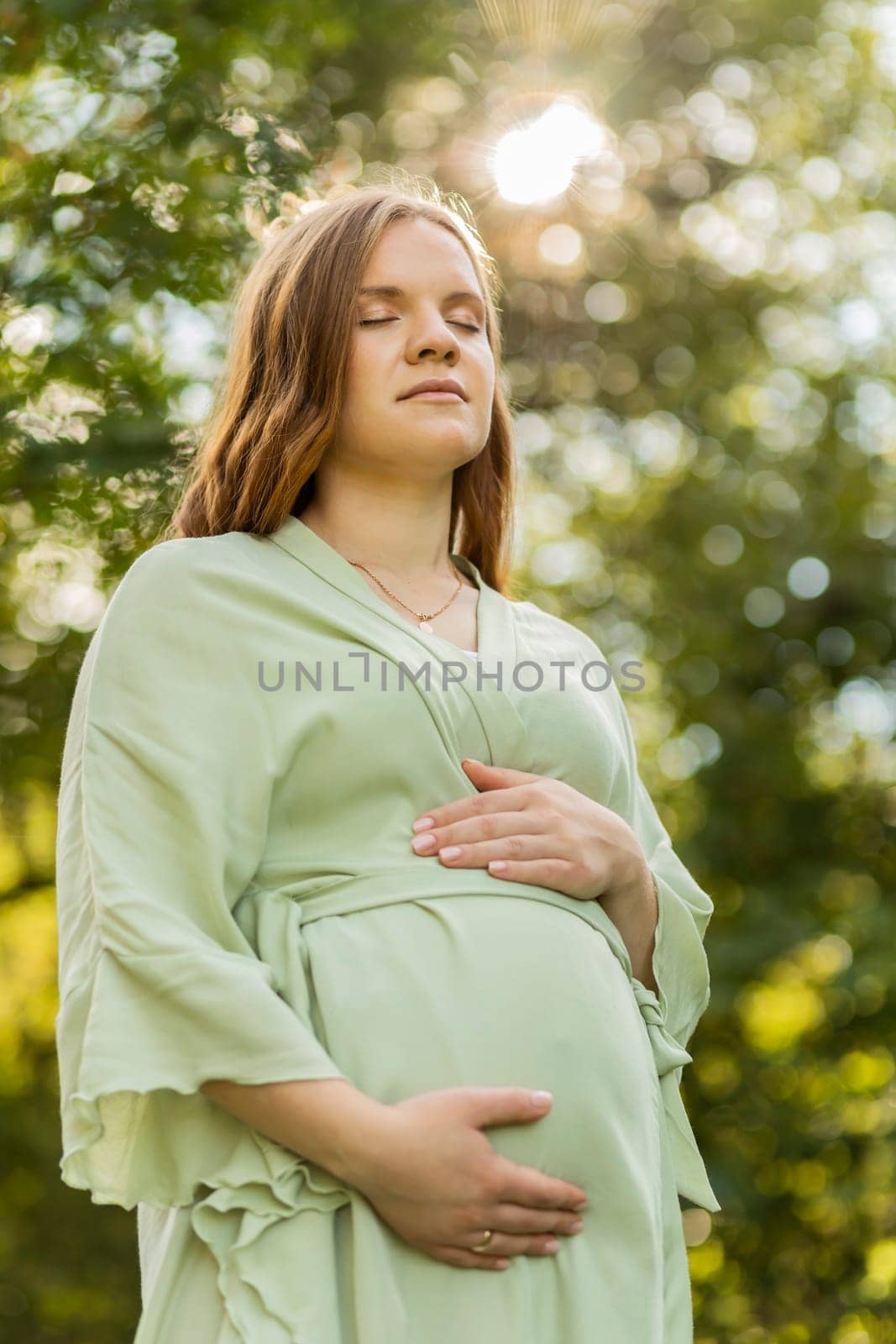 Young pregnant female holds hands on big belly, closed eyes, green trees,sky, sunlight on background. Happy female wears dress. Magic pregnancy. Labor, emotional connection with baby. Vertical plane