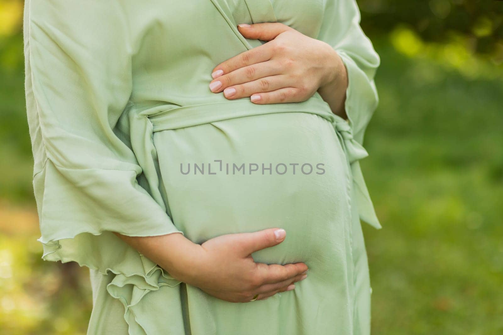 Cropped pregnant woman puts,holds hands on big belly, green trees,meadow on background.White female wears dress.Magic happy pregnancy.Childbirth preparation,emotional connection with baby.Horizontal