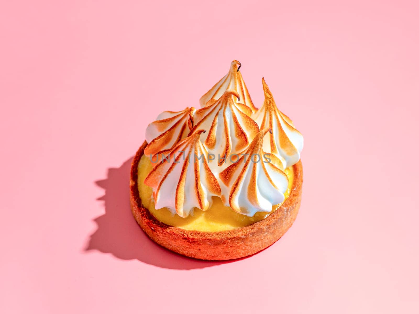 Mini cake decorated with whipped egg whites meringue. Mini lemon tart with burnt French meringue. Delicious dessert with lemon filling. topped with meringue on pink background, hard shadows copy space