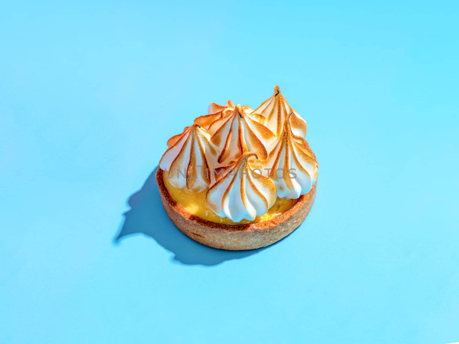 Mini cake decorated with whipped egg whites meringue. Mini lemon tart with burnt French meringue. Delicious dessert with lemon filling. topped with meringue on blue background, hard shadows copy space