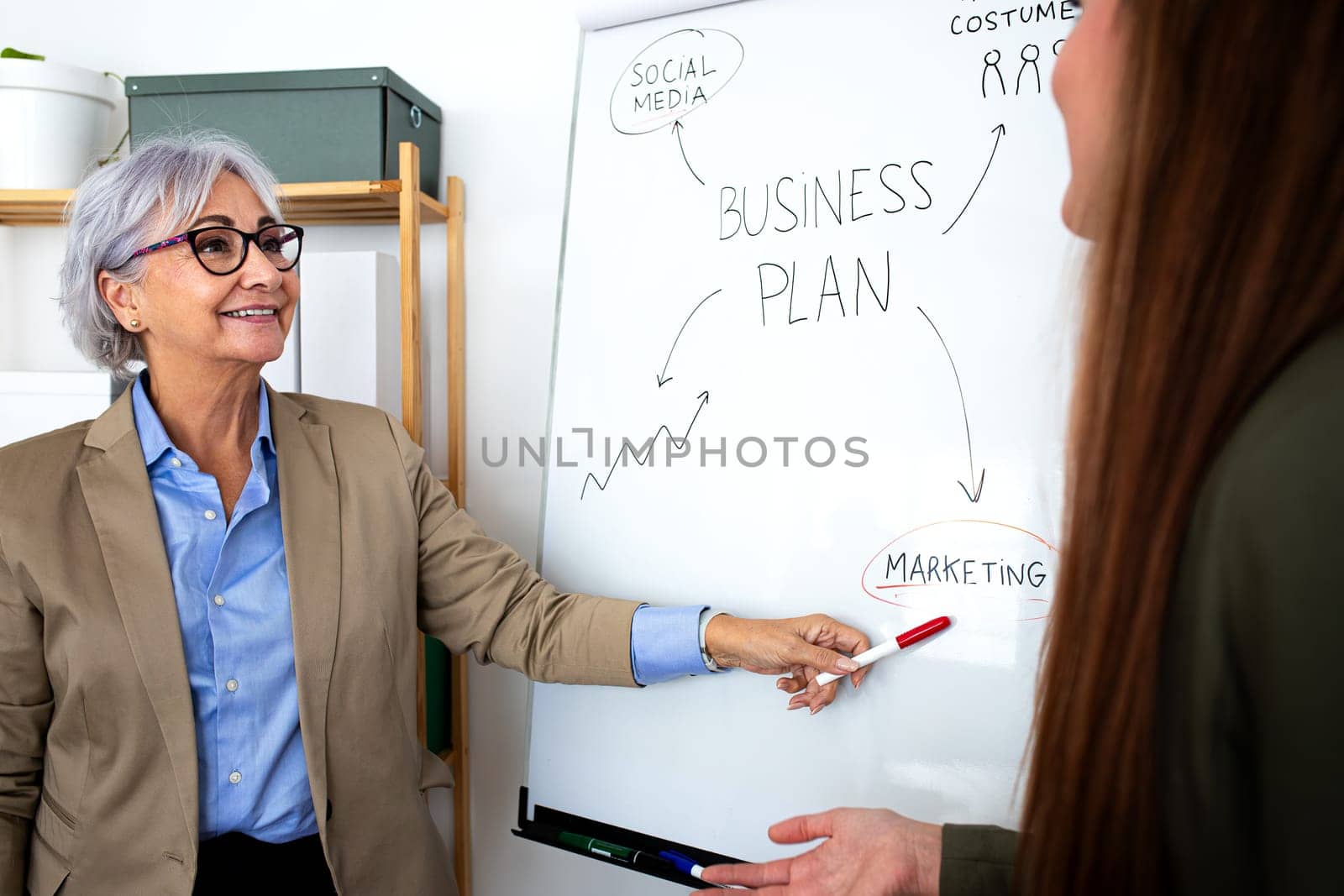 Senior caucasian business woman showing young female coworker business plan on white board in the office. Business concept.