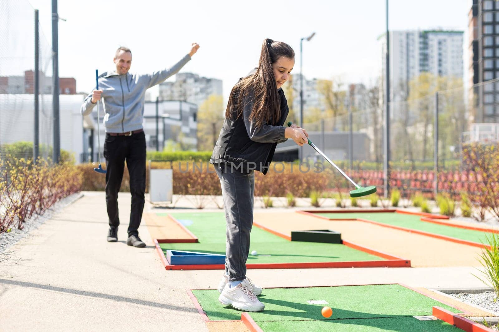 Sharing with golf experience. Cheerful young man teaching his daughter to play mini golf at the day time. Concept of friendly family.
