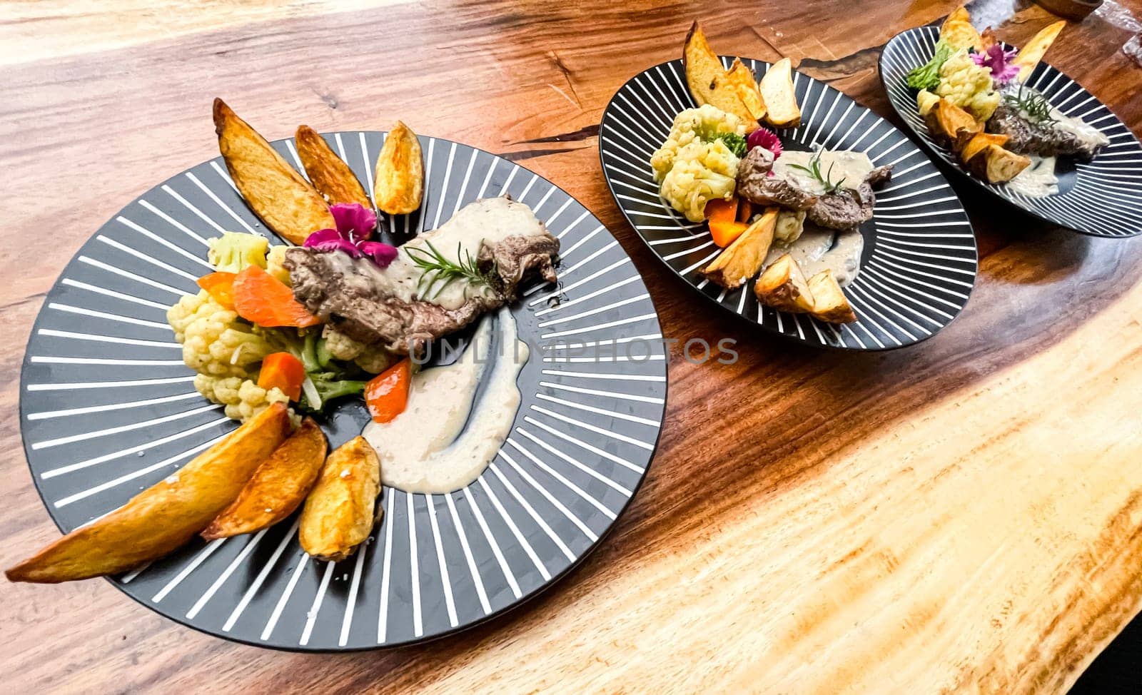 Juicy beef steak with salad and potato wedges along with pepper corn sauce served on black and white stripe on a wooden stone table. Top view
