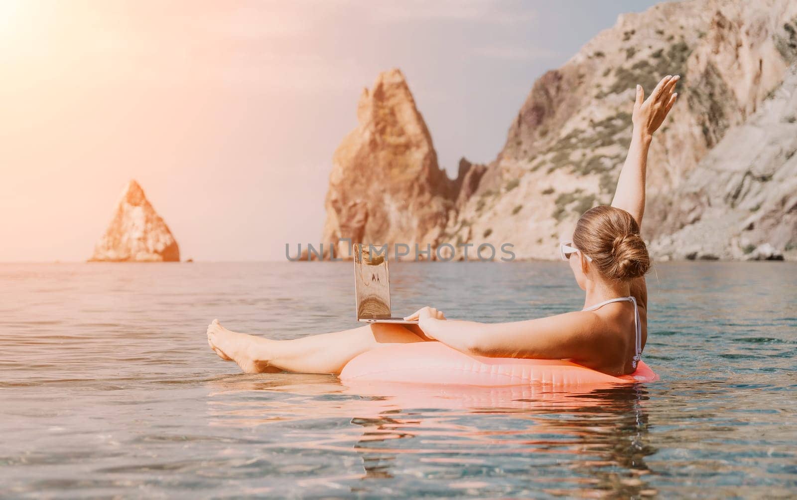 Woman freelancer works on laptop swimming in sea on pink inflatable ring. Pretty lady typing on computer while floating in the sea on inflatable donut at sunset. Freelance, remote work on vacation