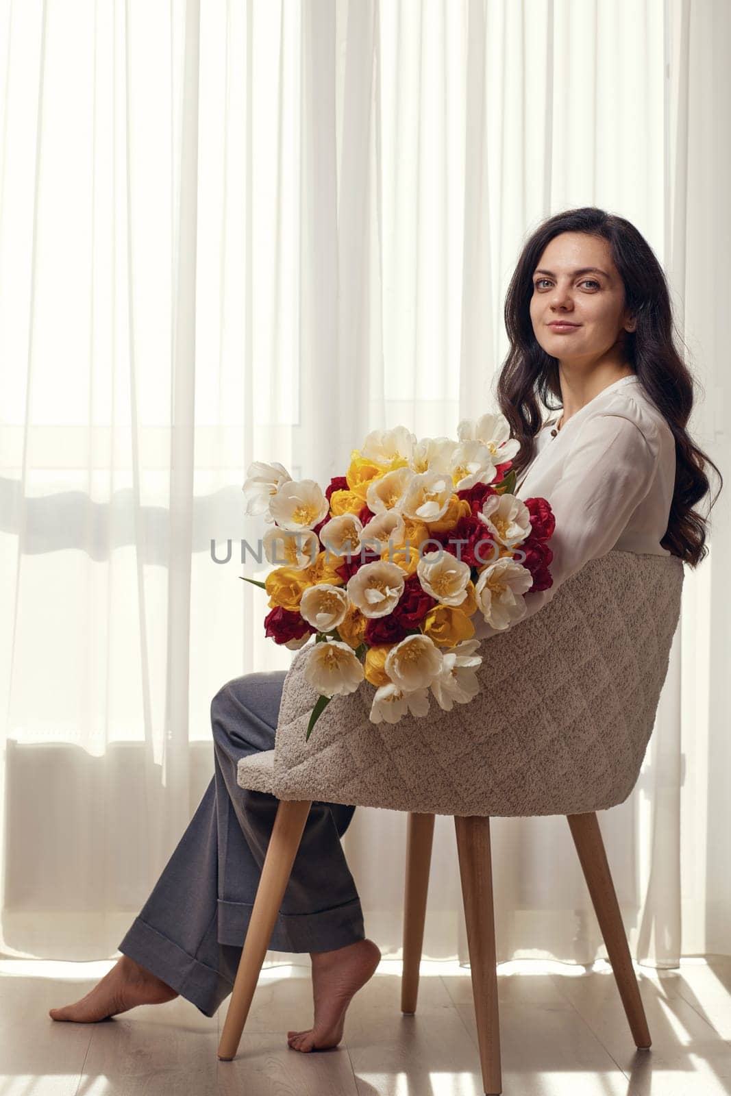 Beautiful woman with spring flowers tulips in hands sitting on chair near window. Women's Day