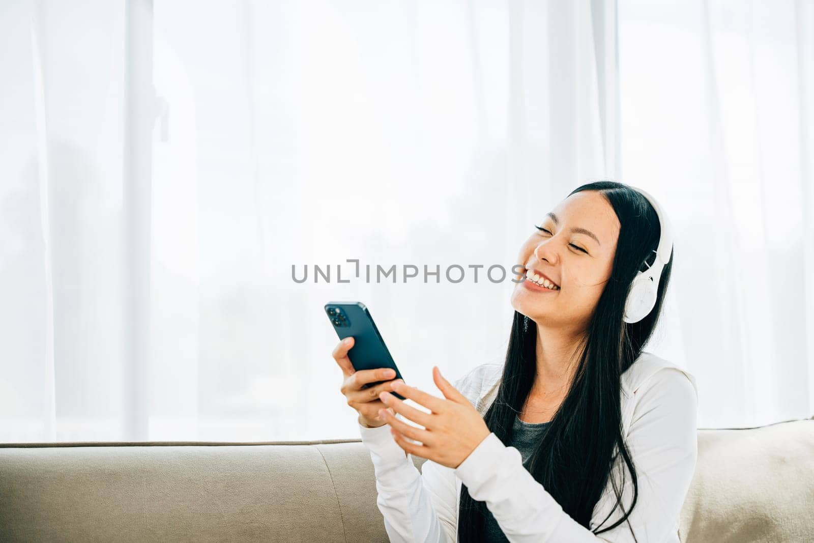 Woman relaxes on sofa listens to music on smartphone wears headphones. Engaged in leisure enjoyment and technology at home. Entertainment and relaxation concept.