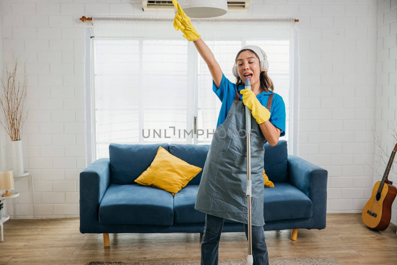 House cleaning turns joyful housewife sings happily while using mop microphone during cleanup. Enjoying domestic chores dancing and cleaning in living room. Cheerful housewife singing playing with mop by Sorapop