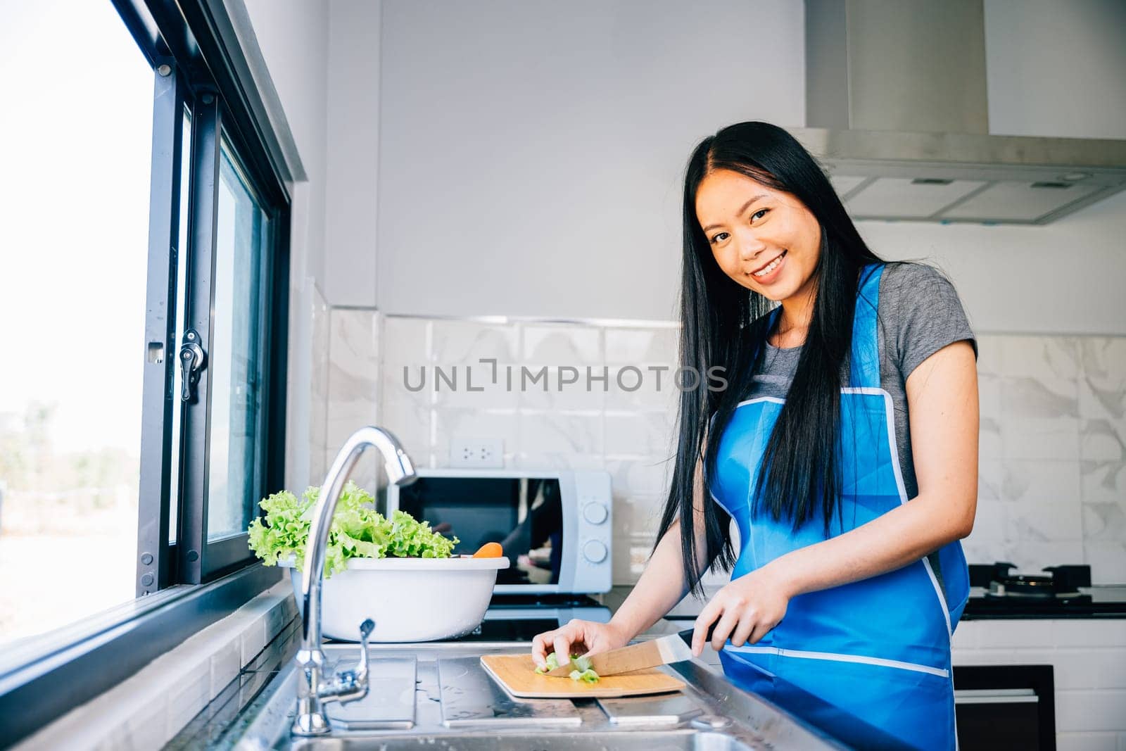 A smiling woman in an apron prepares a healthy dinner cutting vegetables for a delicious salad in her kitchen. Close-up of a cheerful housewife making a nutritious meal.