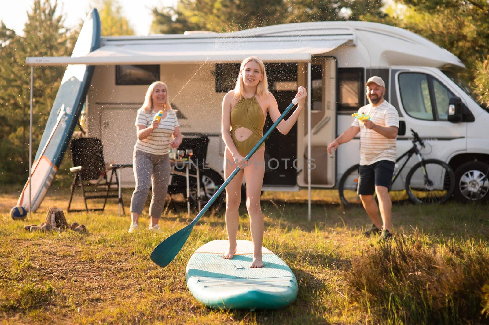 the family is resting next to their mobile home. My daughter is standing with a paddle on a sup board, and her parents pour water on her.