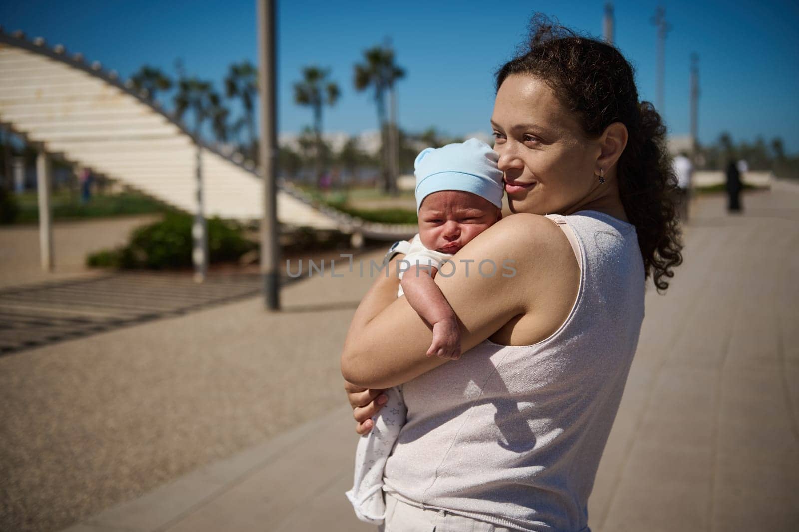 Beautiful delightful young woman. Affectionate loving caring mother gently carrying and embracing her newborn baby, sweetly sleeping in her arms while walking together outdoor