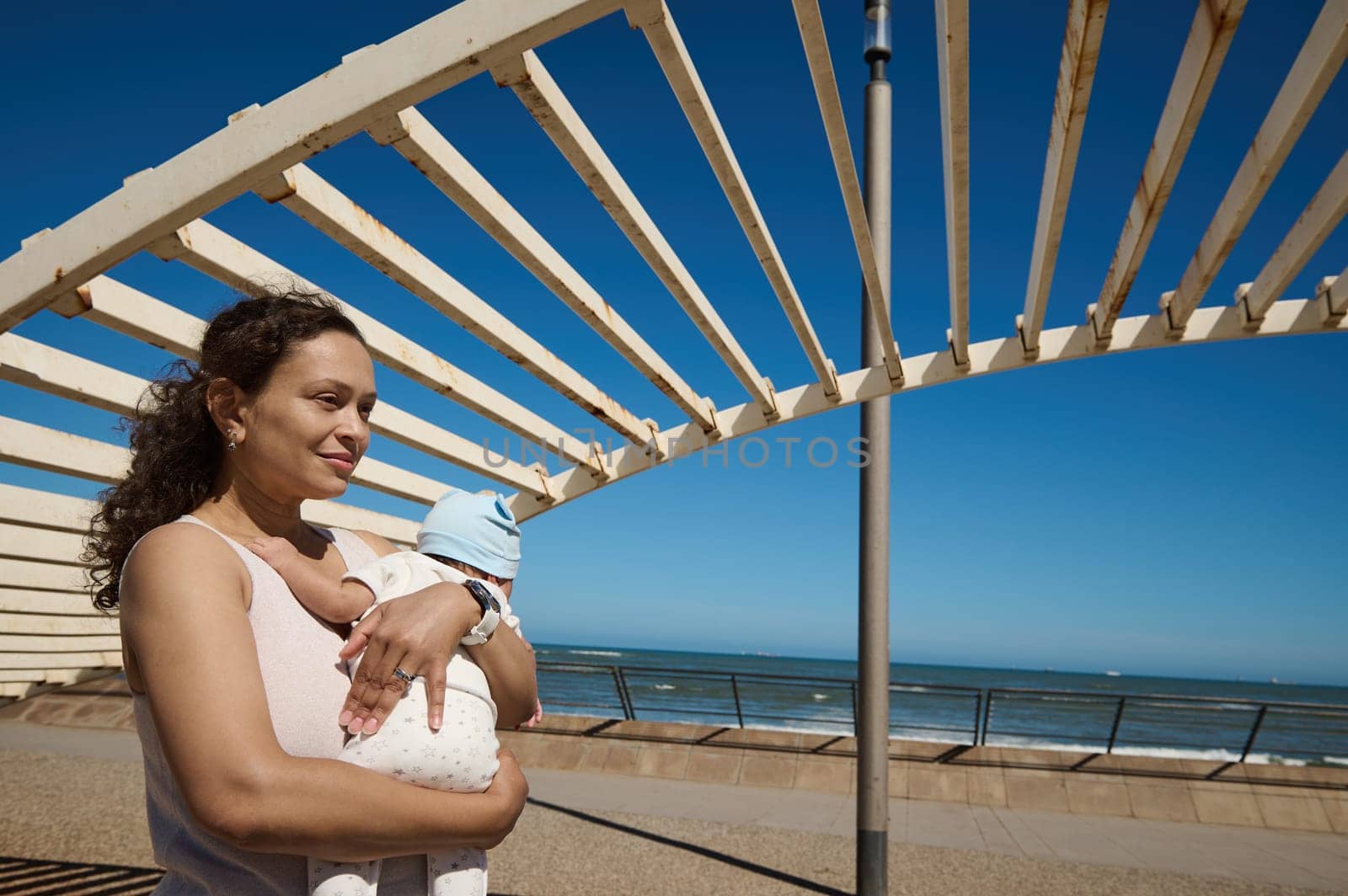 Young smiling pretty woman, mother carrying her newborn baby while walking together on the city on warm sunny day