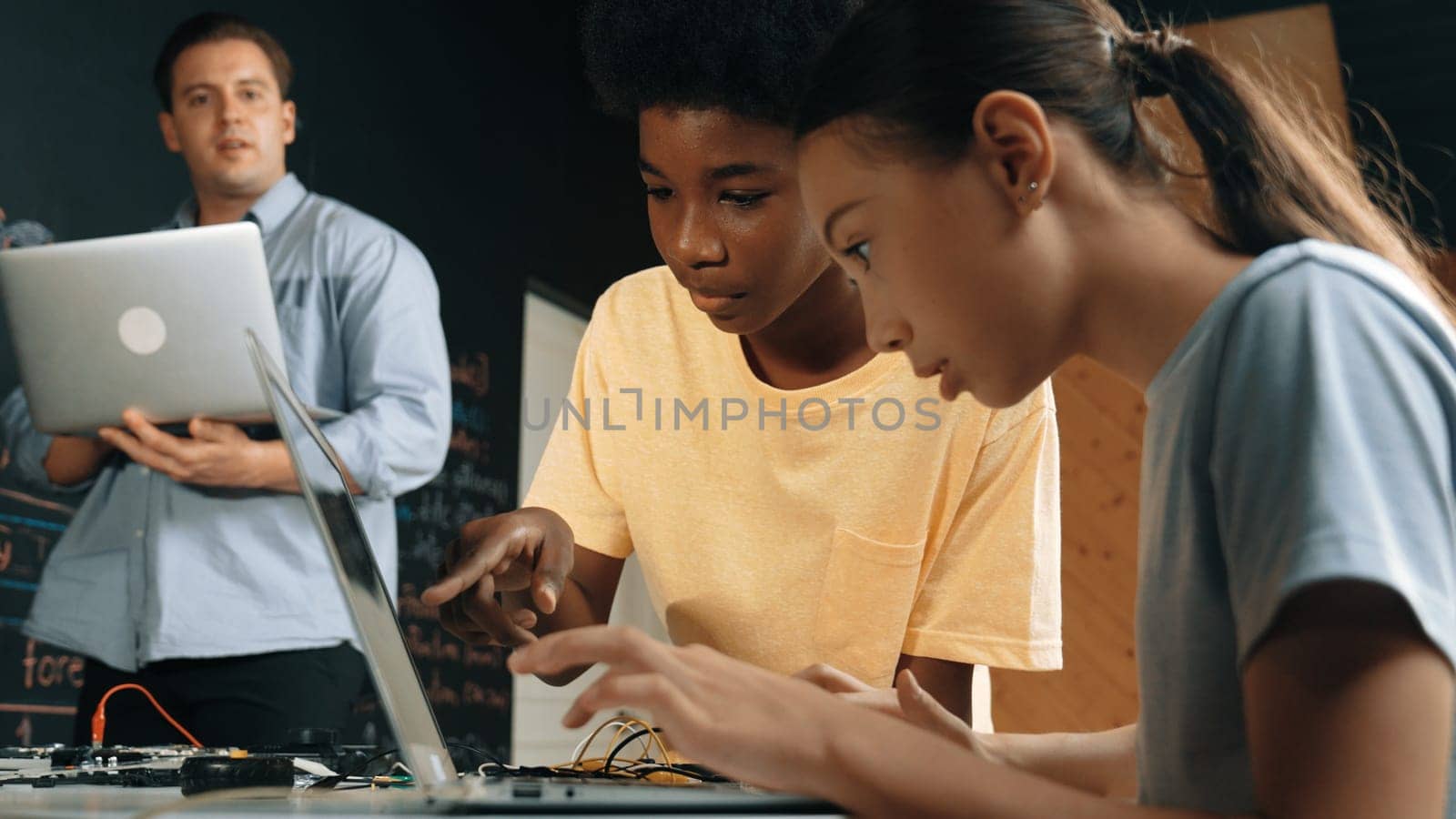 Caucasian girl programing or coding system while african boy fixing model. Teacher holding laptop while looking at diverse children writing web develop program. Innovation education. Edification.