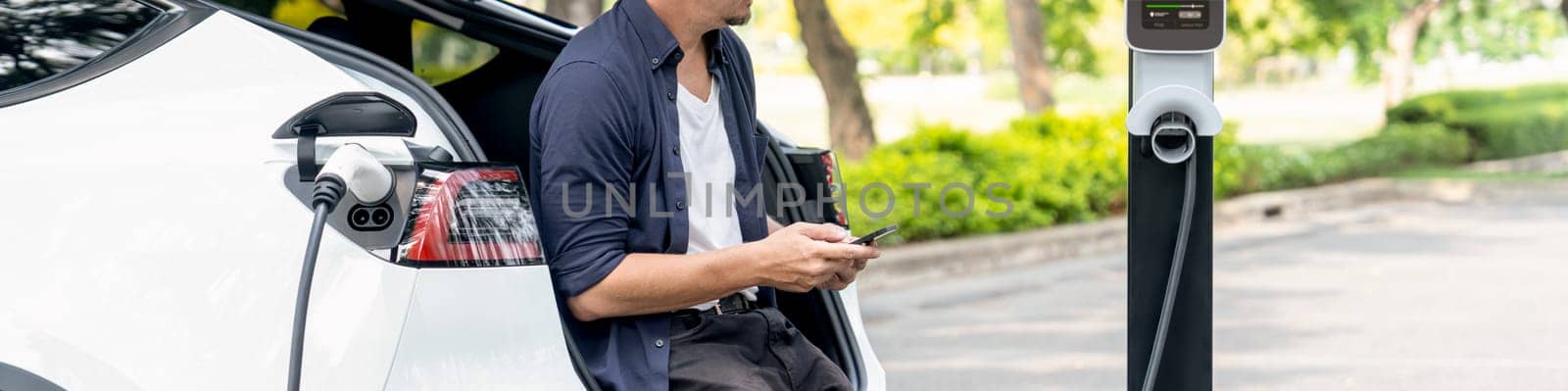 Man using smartphone online banking application to pay for electric car battery charging from EV charging station during vacation road trip at national park or summer forest. Panorama Exalt