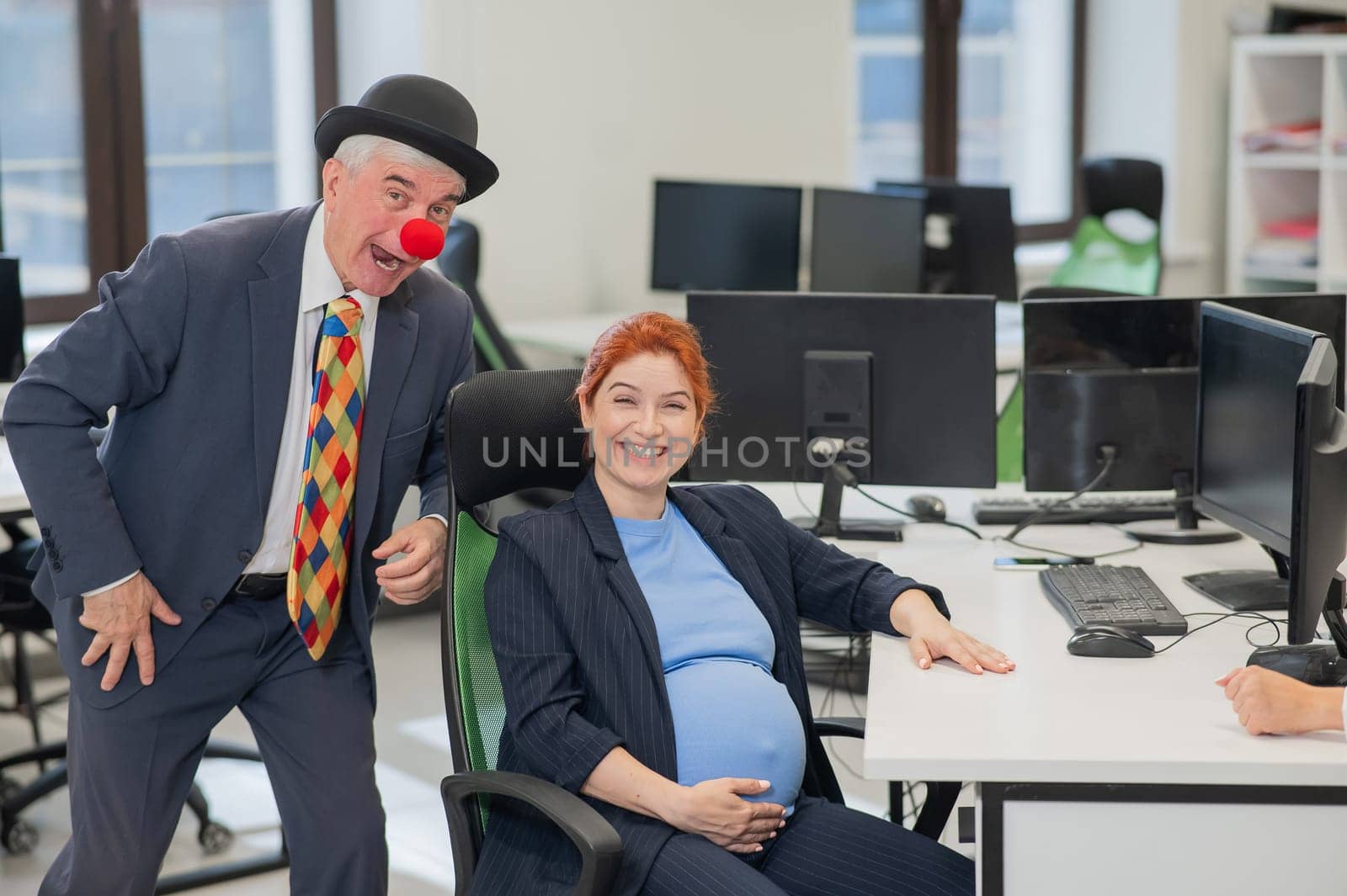 Elderly Caucasian man in clown costume amuses pregnant red-haired woman at work desk in office. by mrwed54