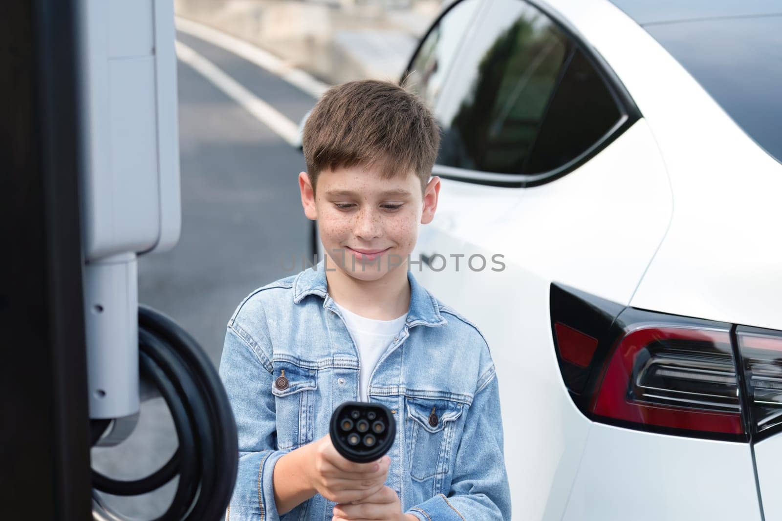 Road trip vacation with eco-friendly EV car and little boy pointing EV charger at camera. Electric vehicle and charging station using clean and sustainable energy for environment protection. Perpetual