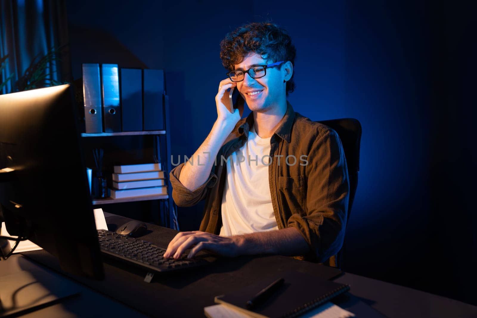 Young businessman calling on smartphone to customer or coworker, looking at computer screen of creative presentation surround work desk at neon modern office. Concept of new next project. Gusher.