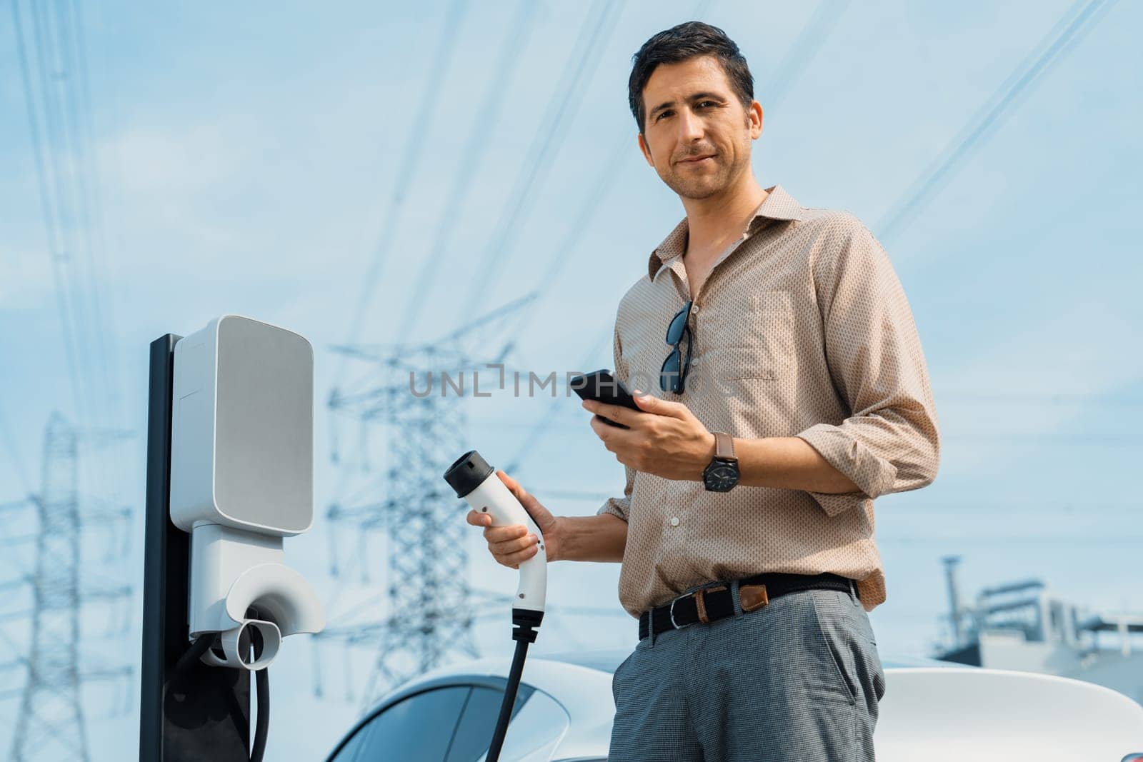 Man pay for electricity with smartphone while recharge EV car battery at charging station connected to power grid tower electrical as electrical industry for eco friendly car utilization.Expedient