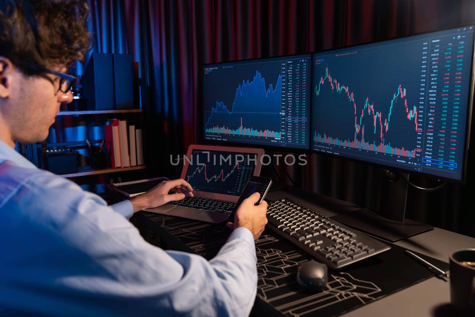Working young business trader focusing on smartphone comparing with laptop in market stock graph in real time screens at neon light modern office. Concept of analyzing dynamic exchange rate. Gusher.