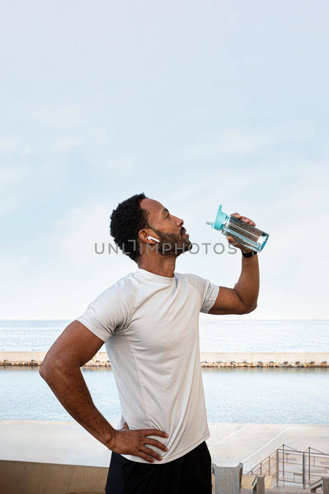 Vertical portrait of Black male runner taking a break to hydrate. African American man drinking water after exercising and jogging outdoors. Copy space. Healthy lifestyle and sport concept.