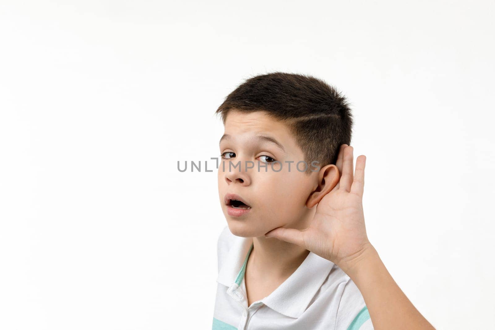 happy little child boy in striped t-shirt making hearing gesture on white background. facial expression. kid with hand over ear listening gossip.