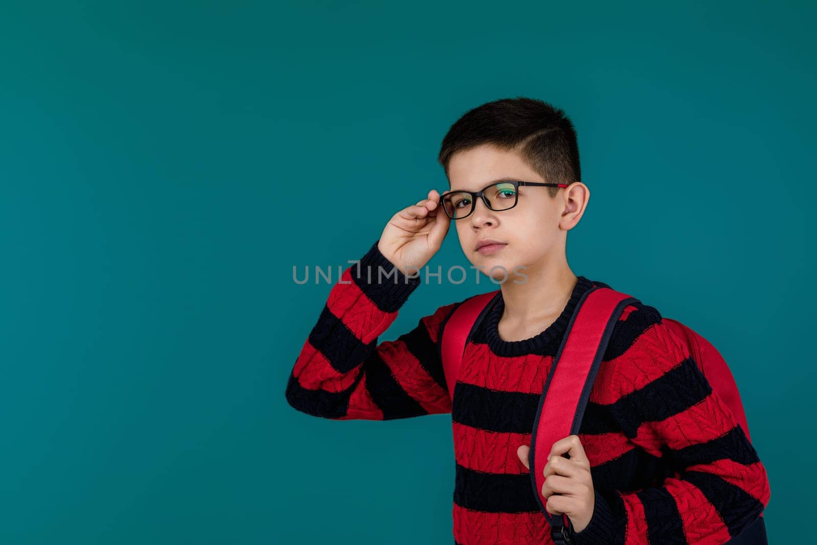 little cheerful school boy wearing glasses with red backpack over blue background. copy space
