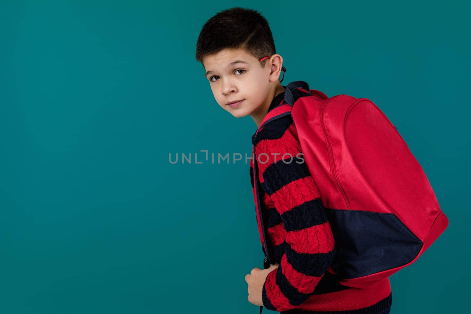 little cheerful school boy with red backpack over blue background. copy space
