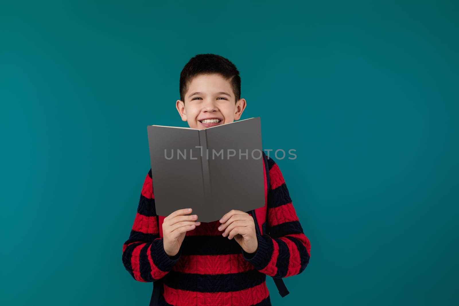 smiling little cheerful school boy reading a book over blue background. child covers his face with book. blank space on book for your text