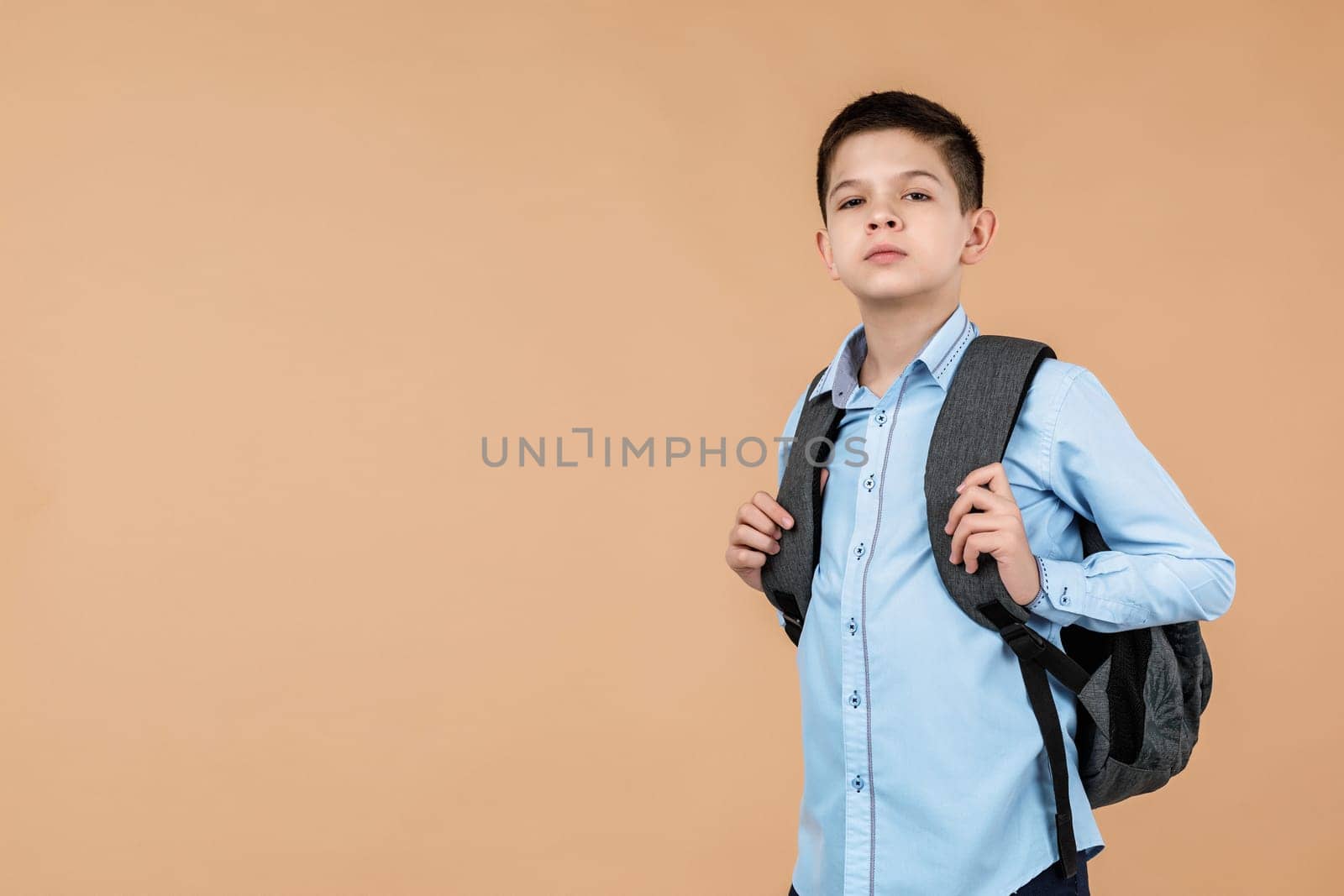 little cute clever school boy with gray backpack looking at the camera over soft yellow background. copy space. Back to school.