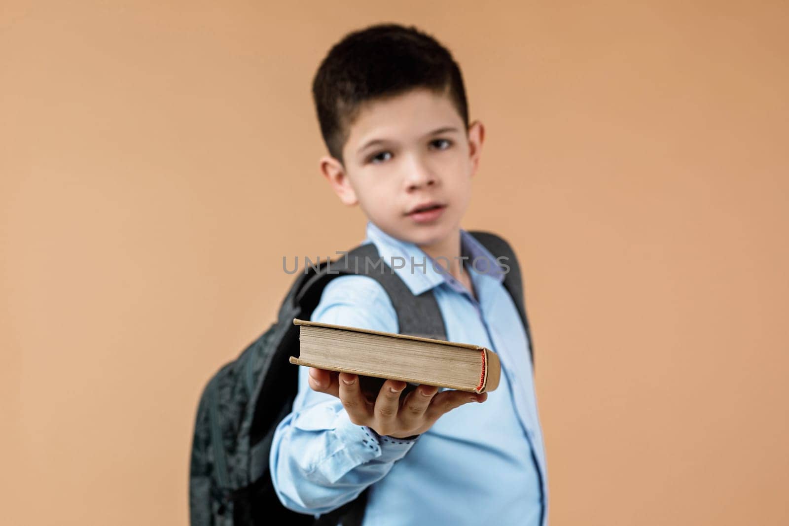 little cheerful school boy with backpack giving book over beige background. focus on book