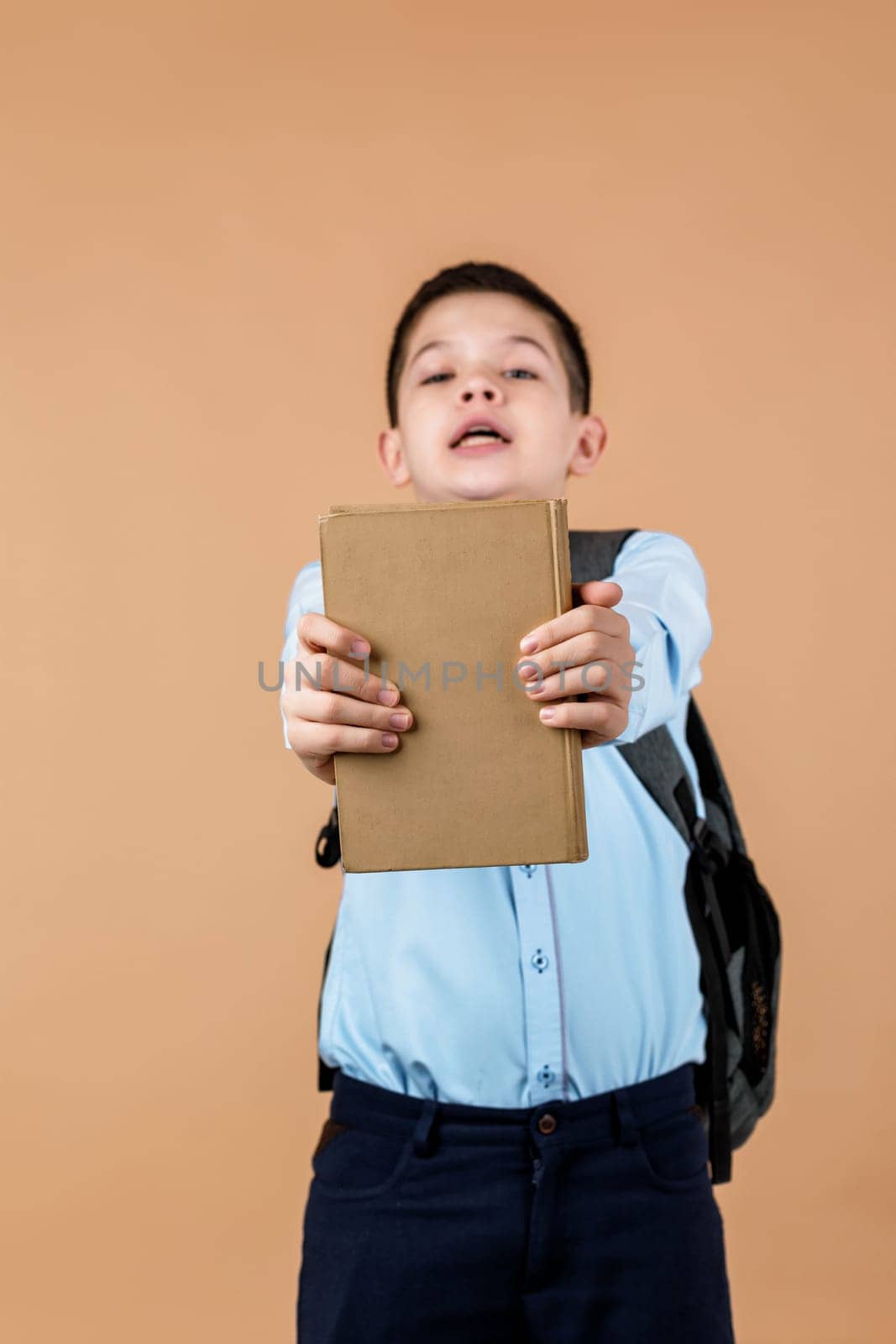 little cheerful school boy with backpack giving book over beige background. focus on book