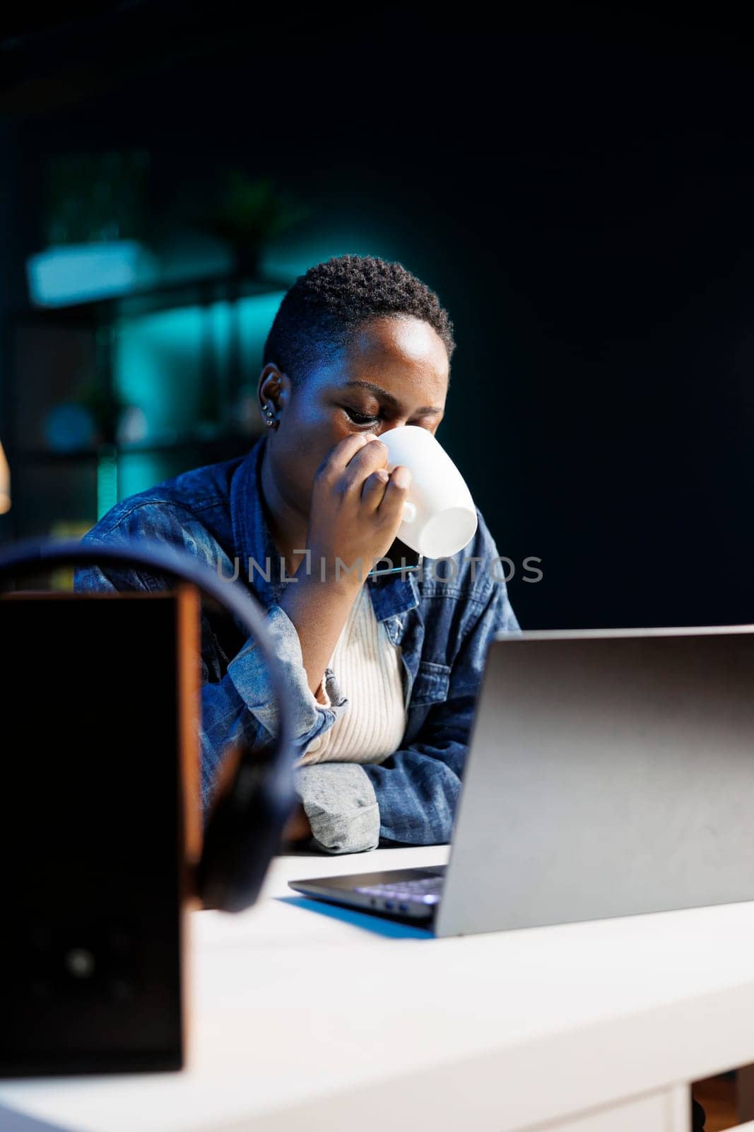 Portrait of african american remote worker drinking coffee while working on her digital laptop. Young black woman with wireless computer on desk is sipping beverage from a cup.