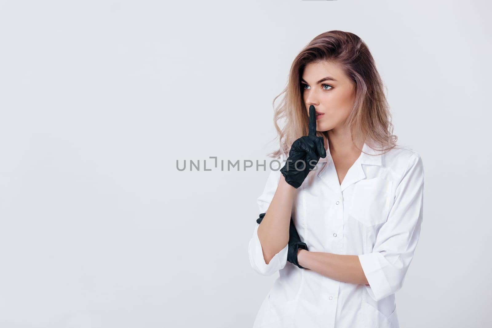 Portrait of young female professional doctor in black gloves shows sign silence. cosmetologist in studio