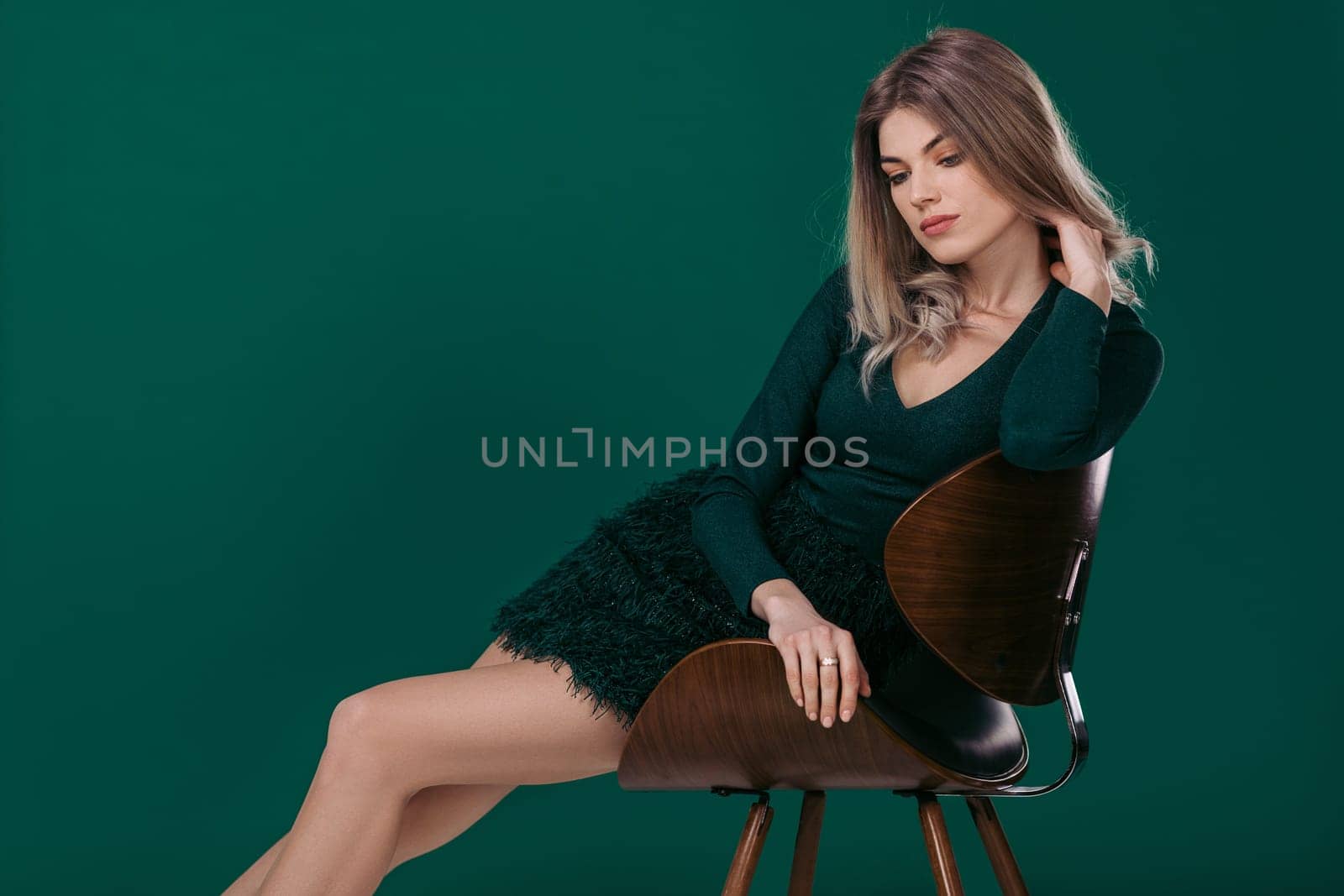 fashion portrait of sensual beautiful blonde woman in green dress sitting on chair against green background. space for text